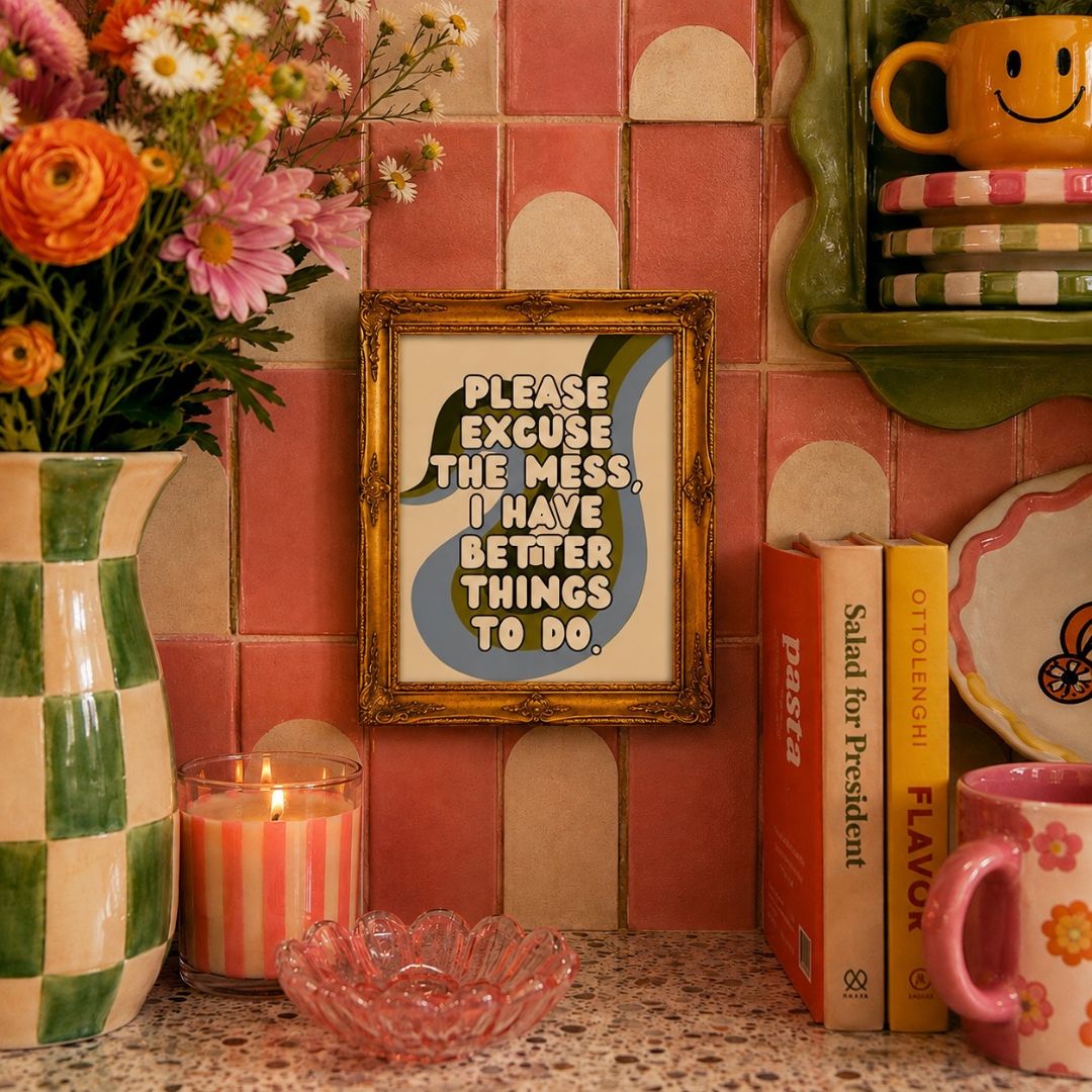 A framed art print with the text 'PLEASE EXCUSE THE MESS, I HAVE BETTER THINGS TO DO.' displayed on a kitchen counter against a pink tiled wall, surrounded by decorative items including a vase of flowers, a candle, books, and a pink mug.