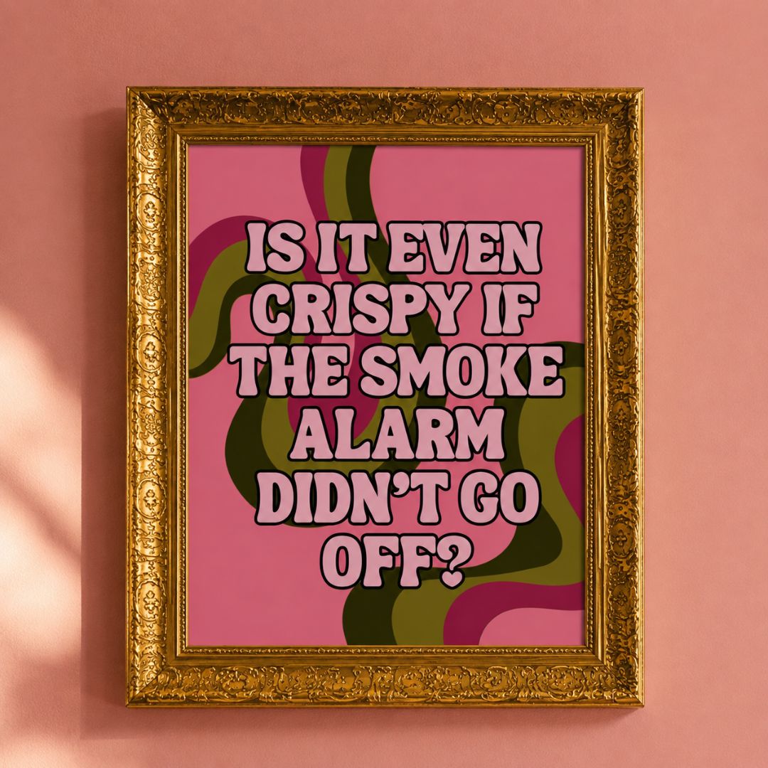 A framed typographic art print hangs on a pink wall. The print has a pink background with retro wavy green and purple lines and bold pink text reading, 'IS IT EVEN CRISPY IF THE SMOKE ALARM DIDN'T GO OFF?' The frame is ornate gold.