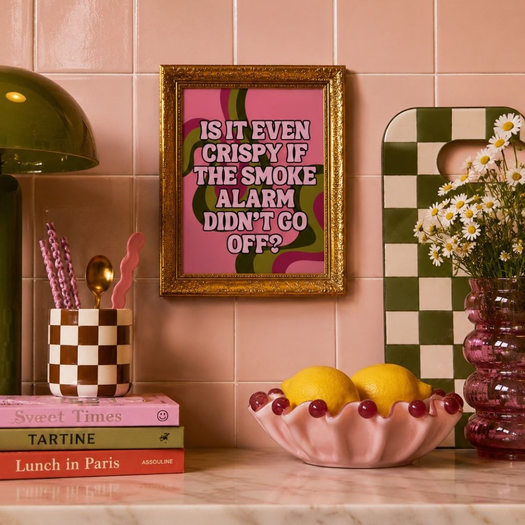 A framed retro-style typographic wall art print with pink, green, and white colors and a gold frame, displayed on a pink tiled kitchen wall above a marble countertop with decorative items including lemons, a pink bowl, a checkered cup, and a green-and-white checkerboard cutting board.