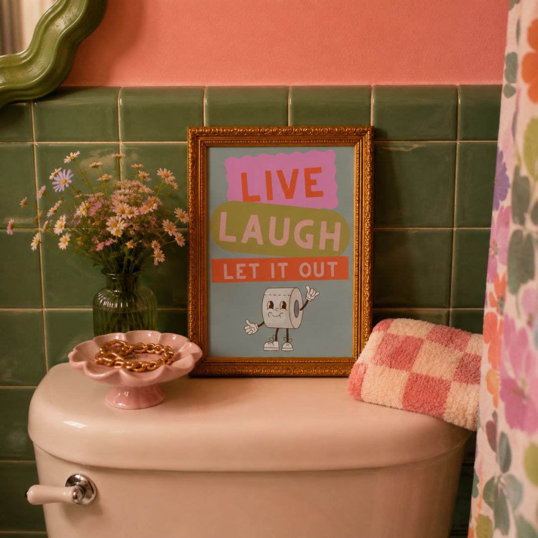 A framed art print with the text 'LIVE LAUGH LET IT OUT' and a cartoon toilet paper character sits on a pink bathroom toilet. The scene includes a pink towel, a small pink dish with gold coins, a green tiled wall, and a pink wall in the background.