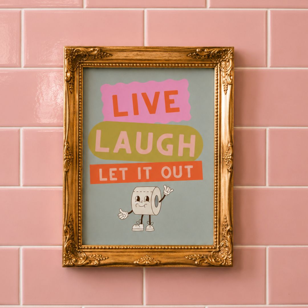 A framed typographic wall art print hangs on a pink tiled wall. The artwork has the words “LIVE LAUGH LET IT OUT” in colorful blocks above a cartoon toilet paper roll character.