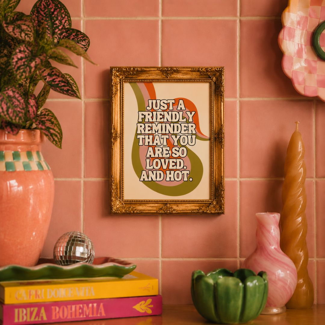 A framed retro-style quote print hangs on a pink tiled wall above a shelf with decorative objects. The print has a gold frame and reads, 'JUST A FRIENDLY REMINDER THAT YOU ARE SO LOVED, AND HOT.