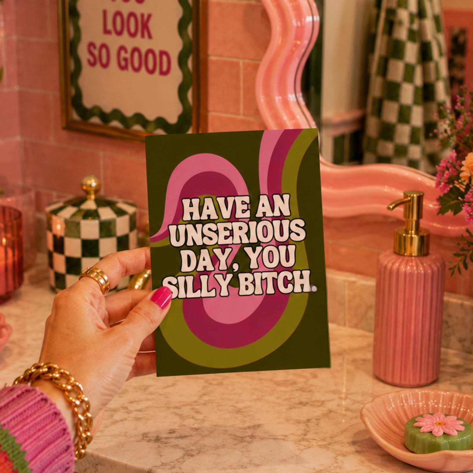 A hand holding a square greeting card with a dark green background and bold pink and olive-green abstract shapes, featuring the text 'HAVE AN UNSERIOUS DAY, YOU SILLY BITCH.' in white.