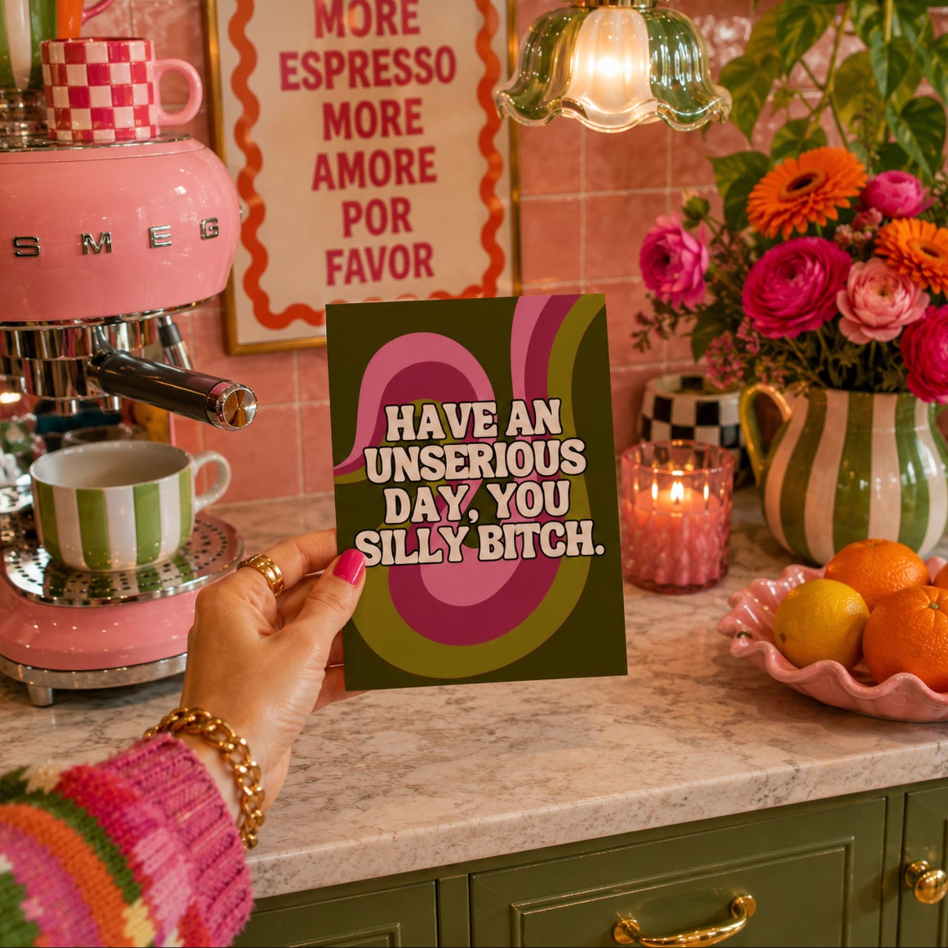 A hand holds a retro-style novelty card with the text 'HAVE AN UNSERIOUS DAY, YOU SILLY BITCH.' in a colorful kitchen setting with a pink espresso machine, flowers, and a sign that reads 'MORE ESPRESSO MORE AMORE POR FAVOR'.