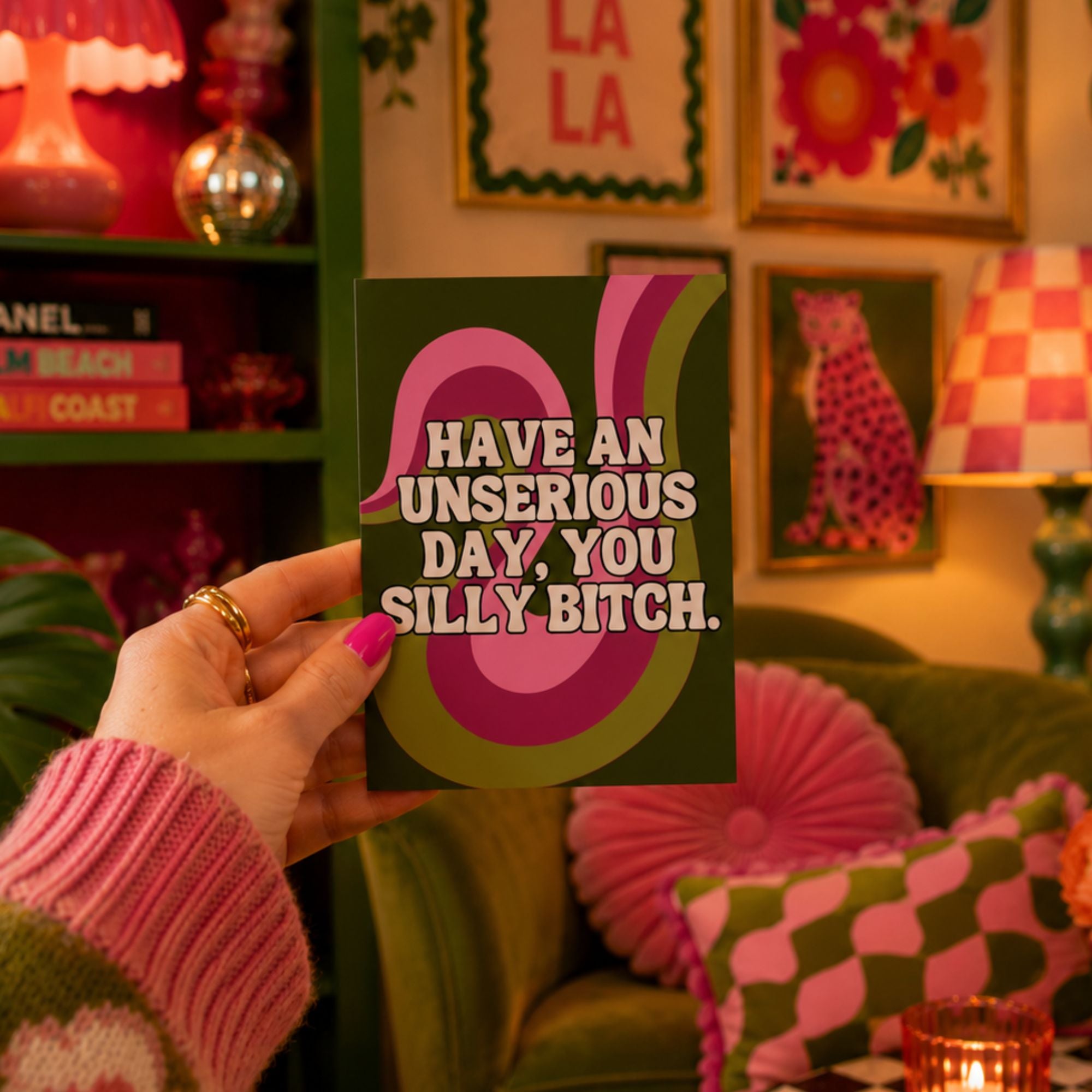 A hand holding a dark green greeting card with bold pink and olive-green retro swirls and the text 'HAVE AN UNSERIOUS DAY, YOU SILLY BITCH.' in white.