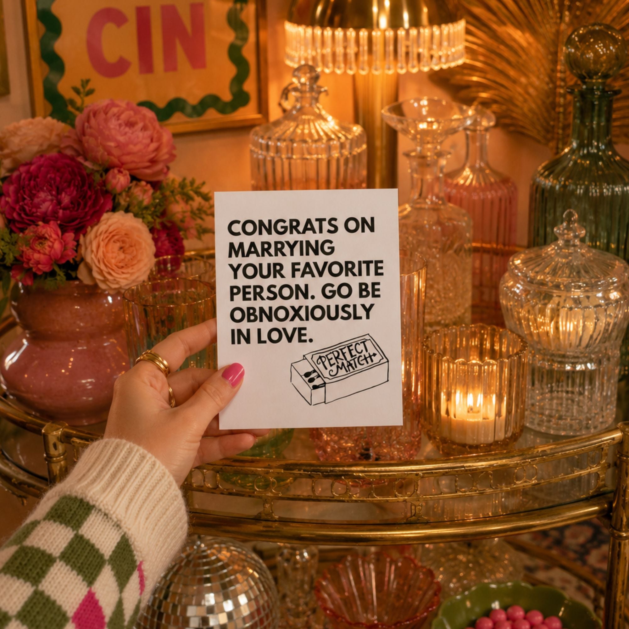 A hand holds a white greeting card that reads 'CONGRATS ON MARRYING YOUR FAVORITE PERSON, GO BE OBNOXIOUSLY IN LOVE.' with a small 'Crazy Exes Cartoon' sketch.