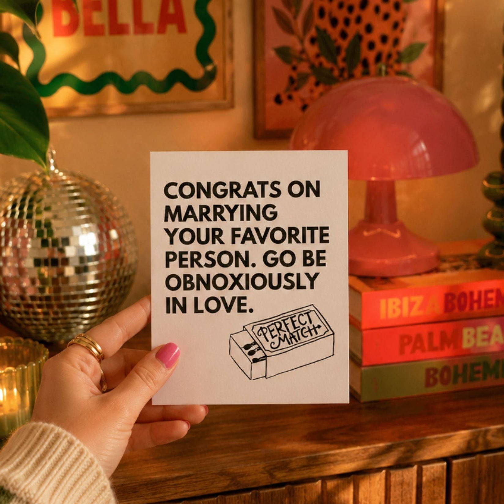 A hand holds a white greeting card with black text that reads 'CONGRATS ON MARRYING YOUR FAVORITE PERSON. GO BE OBNOINOUSLY IN LOVE.