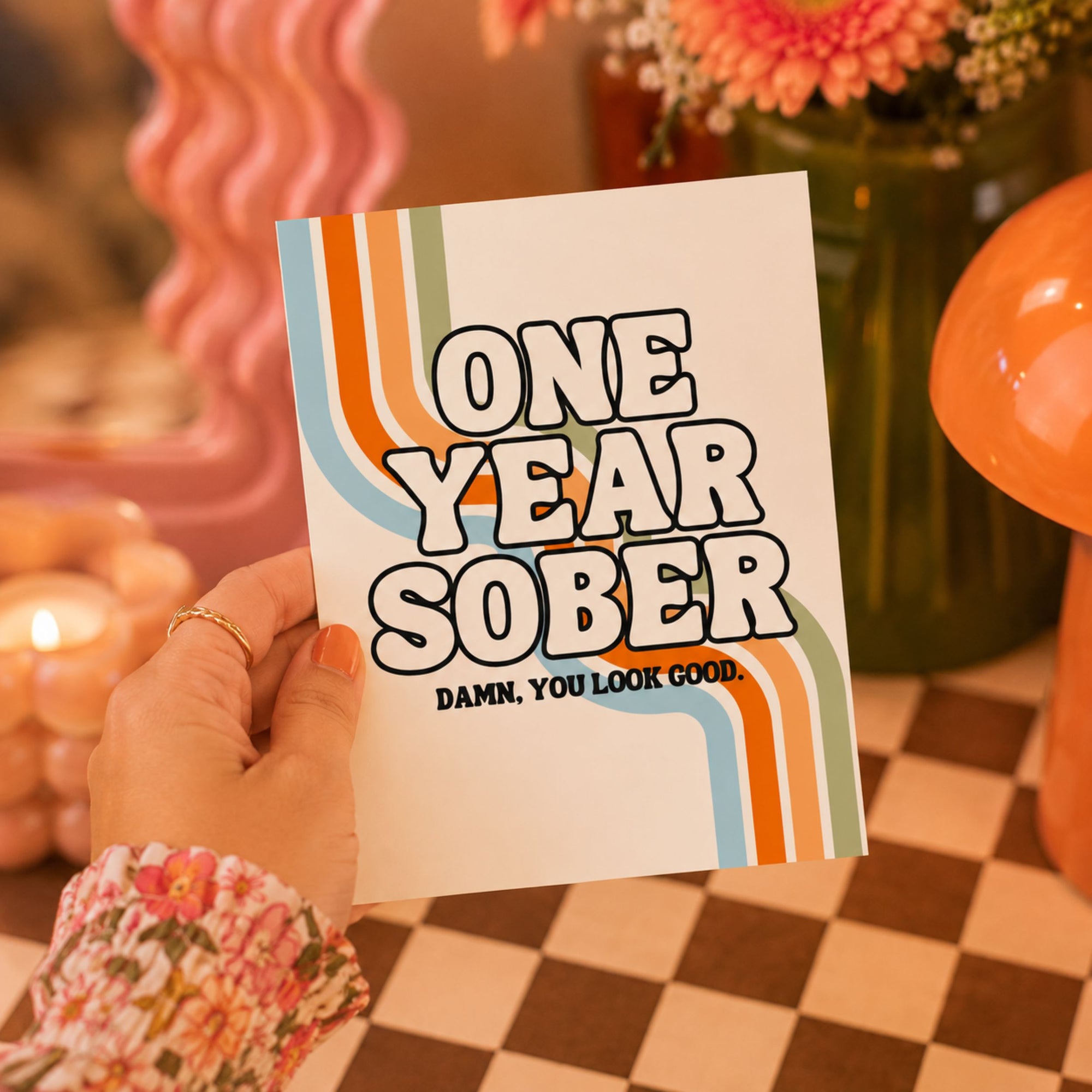 A hand holds a white card with retro-style orange, blue, and beige wavy stripes and the text 'ONE YEAR SOBER' and 'DAMN, YOU LOOK GOOD.' The background is a warm, boho-style setting with a checkered table, flowers, and pink decor.