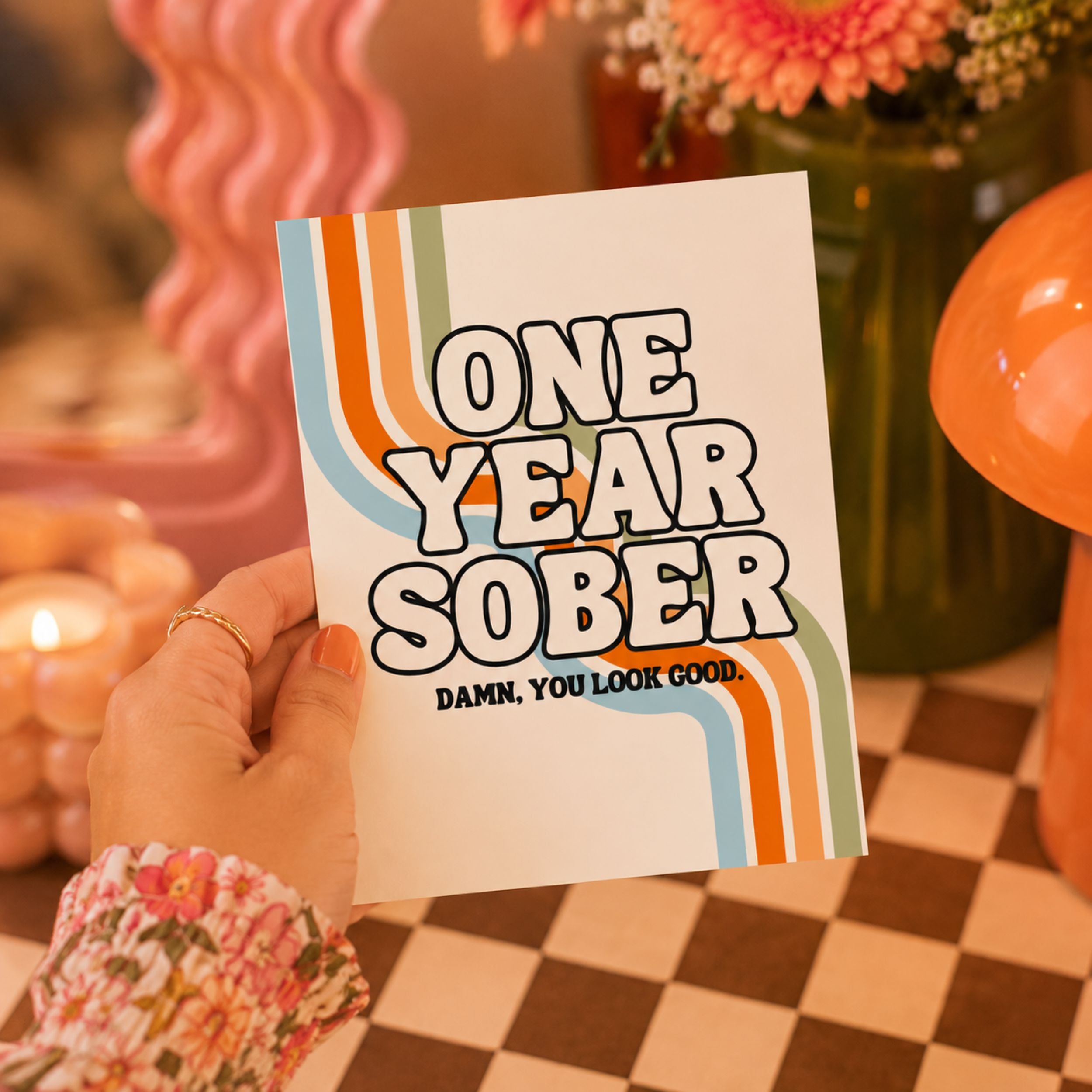 A hand holds a white card with retro-style orange, blue, and beige wavy stripes and the text 'ONE YEAR SOBER' and 'DAMN, YOU LOOK GOOD.' The background is a warm, boho-style setting with a checkered table, flowers, and pink decor.
