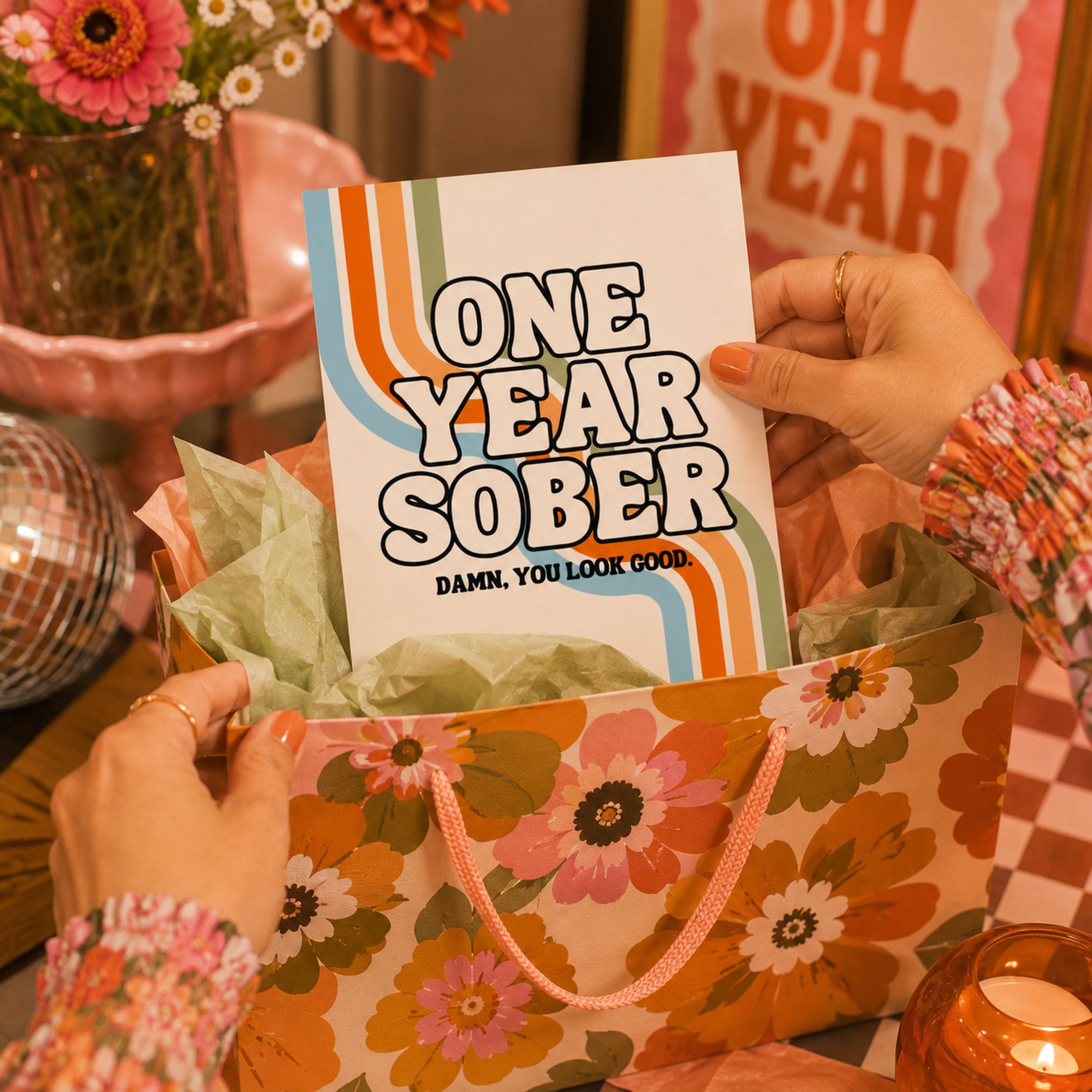 A person holds a white greeting card that reads 'ONE YEAR SOBER DAMN, YOU LOOK GOOD.' in bold black letters with retro orange and blue stripes.