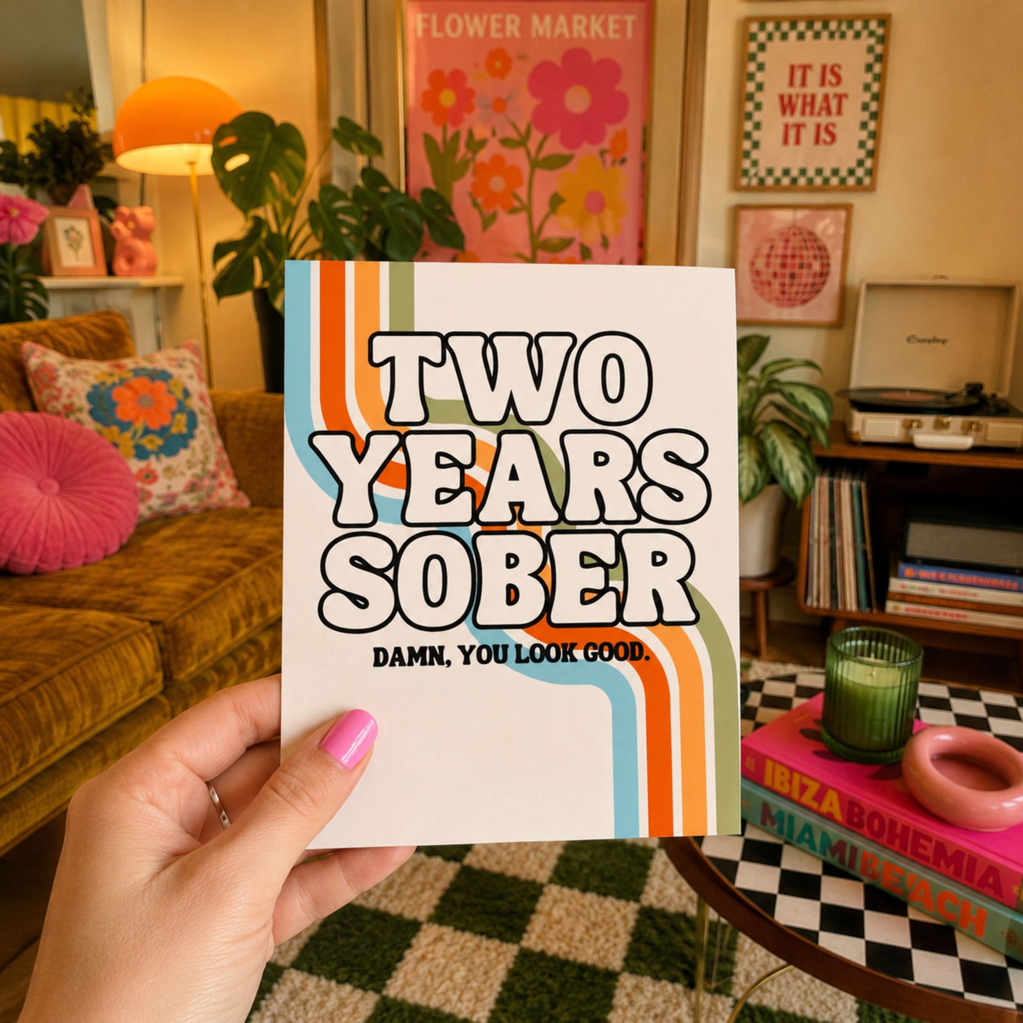 A hand holds a white square card or book cover in a cozy, decorated living room. The front reads 'TWO YEARS SOBER' in bold black letters with retro orange and light blue stripes, and smaller text below says 'DAMN, YOU LOOK GOOD.