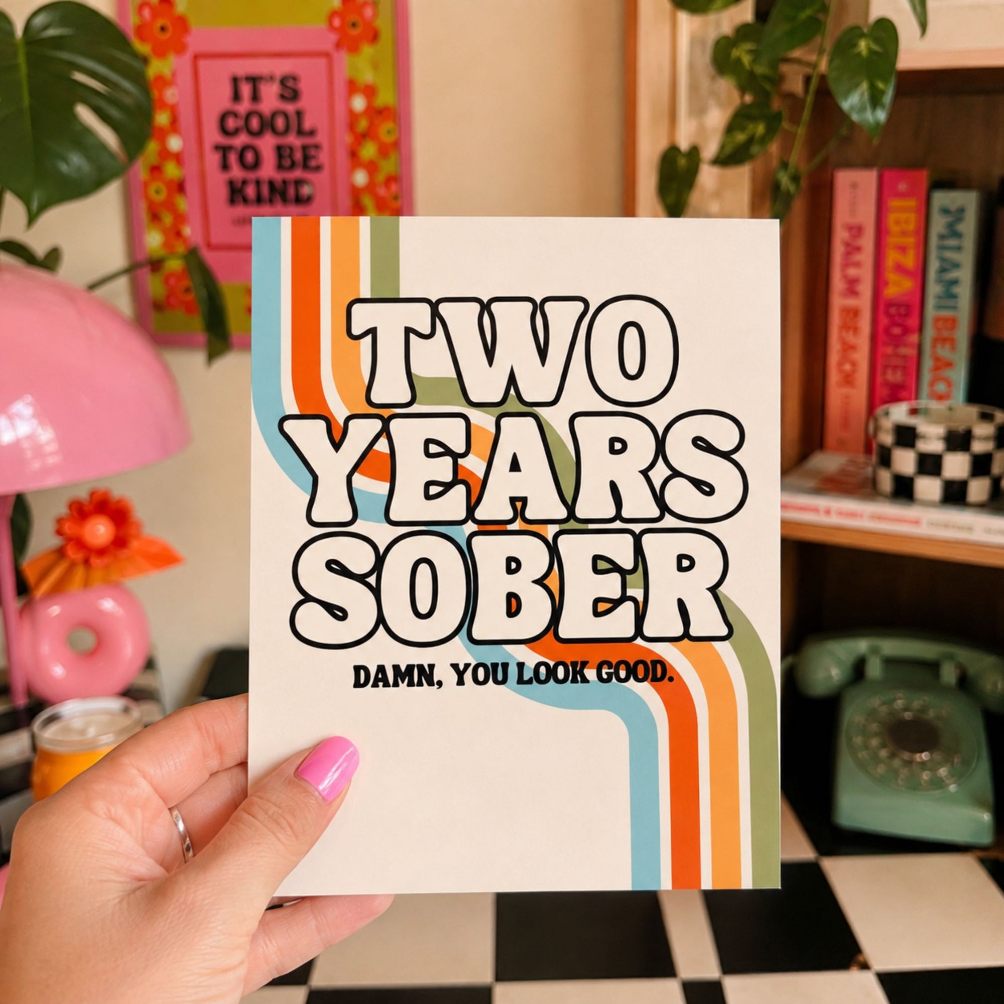 A hand with pink nail polish holds a white greeting card that reads 'TWO YEARS SOBER' with retro orange, blue, and green wavy stripes, and the subtitle 'DAMN, YOU LOOK GOOD.' The background shows a cozy, decorated room with a pink lamp, books, and a retro green rotary phone.