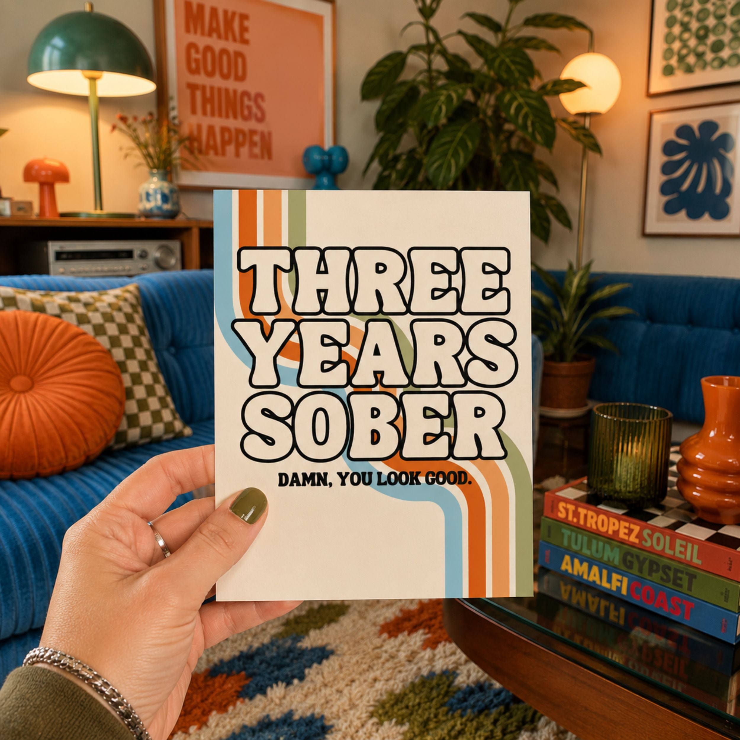 A hand holds a white card or book cover with retro-style orange and blue stripes and the text 'THREE YEARS SOBER' and 'DAMN, YOU LOOK GOOD.' in a cozy living room setting with a blue sofa, plants, and decor in the background.