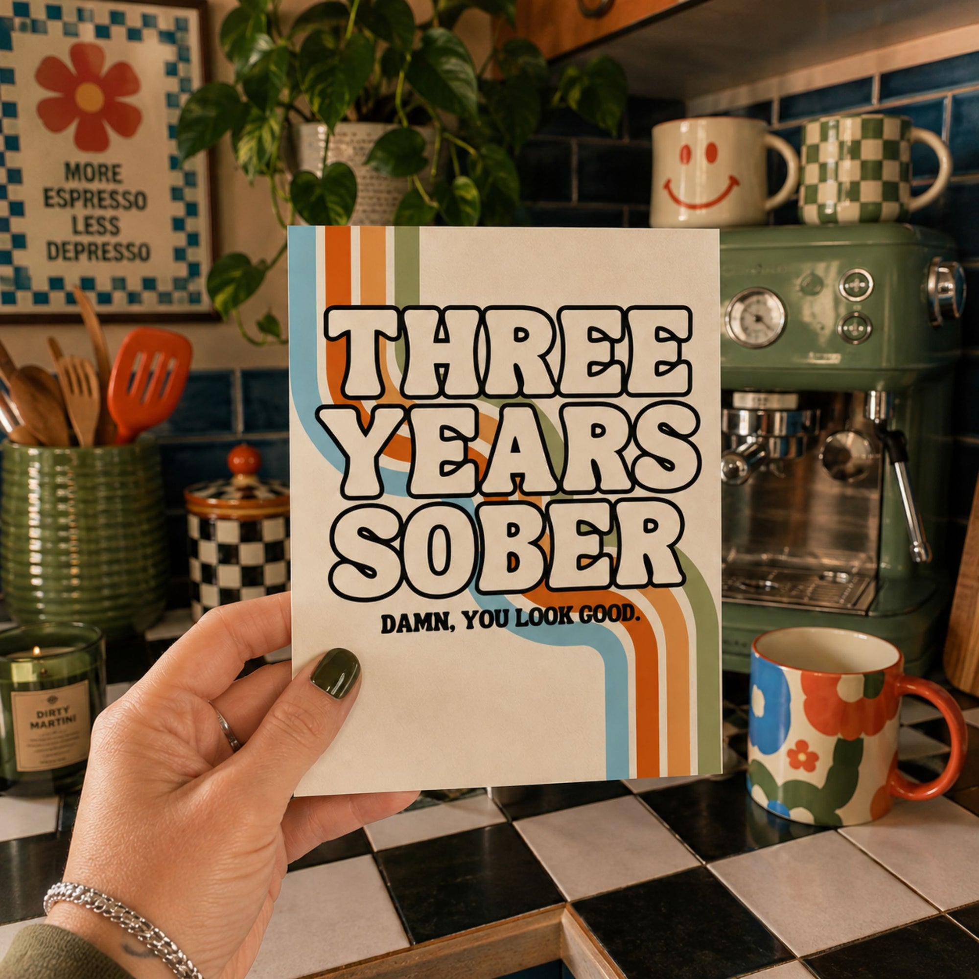 A hand holds a square card or print that reads 'THREE YEARS SOBER' with retro orange, blue, and cream stripes, against a cozy kitchen background with mugs, a plant, and a green espresso machine.