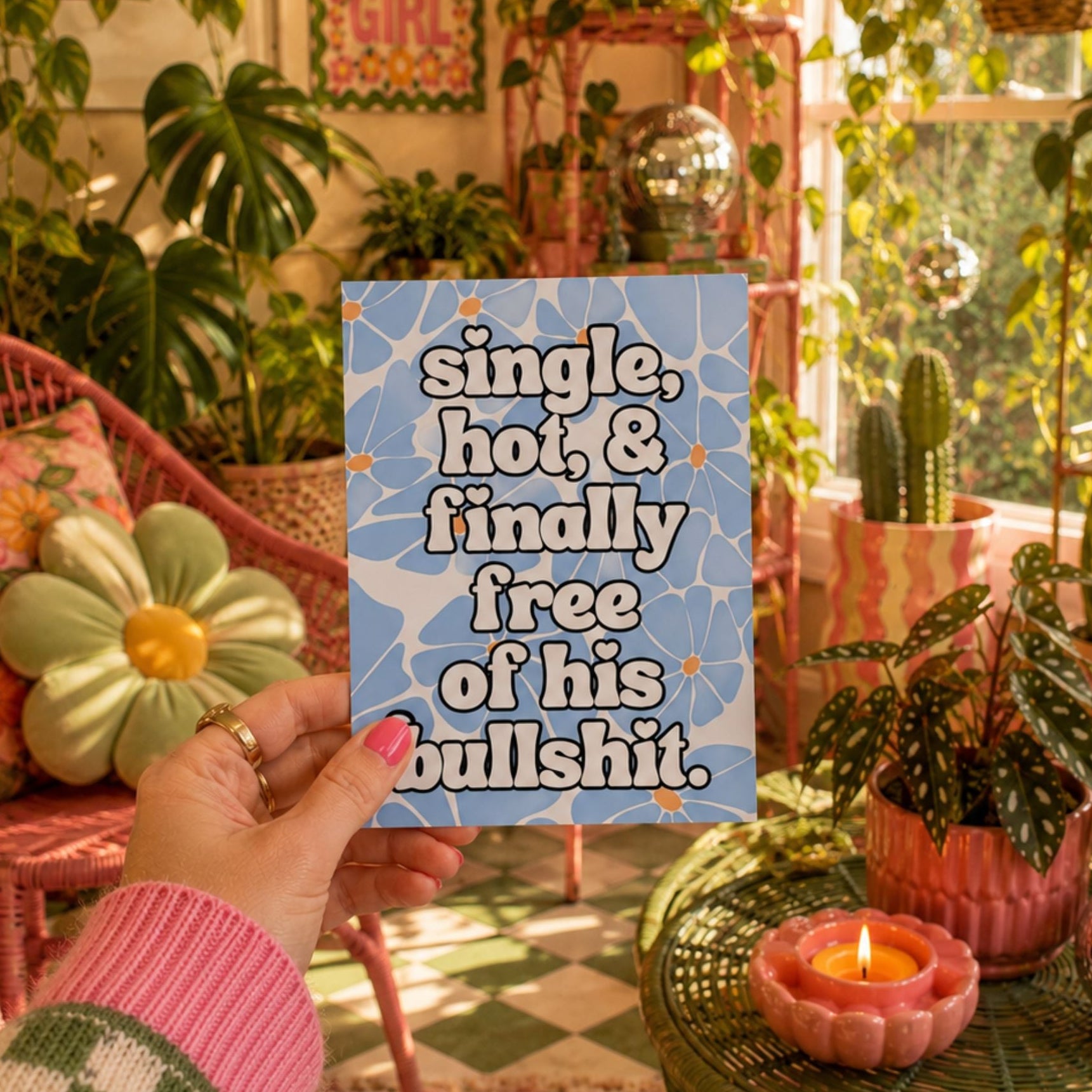 A hand holds a square blue sign with white text reading 'single, hot, & finally free of his bullshit' against a cozy, sunlit plant-filled room with pink furniture and candles.