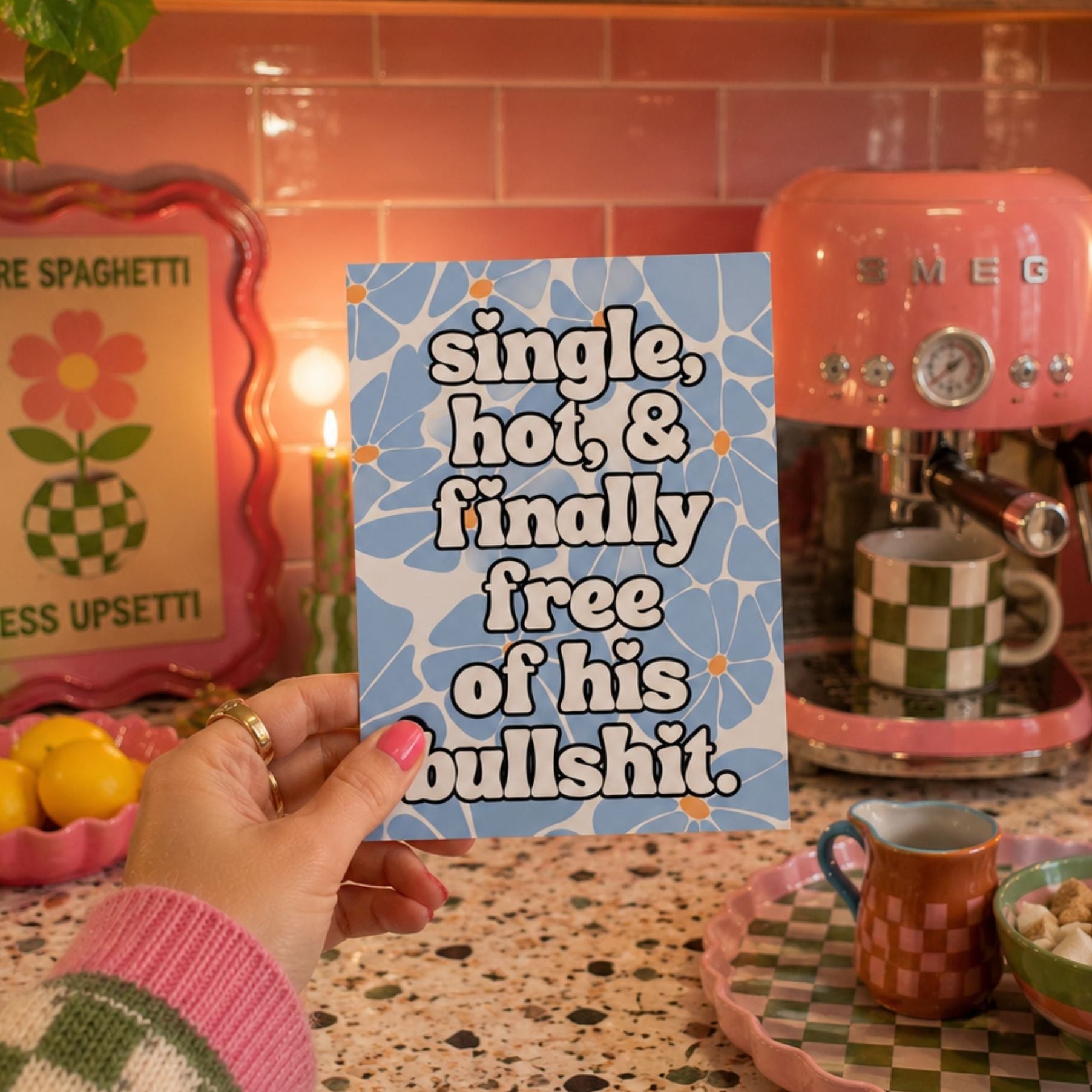 A hand holds a square blue card with white text reading 'single, hot, & finally free of his bullshit' against a retro pink kitchen scene with a pink espresso machine and checkered cups.