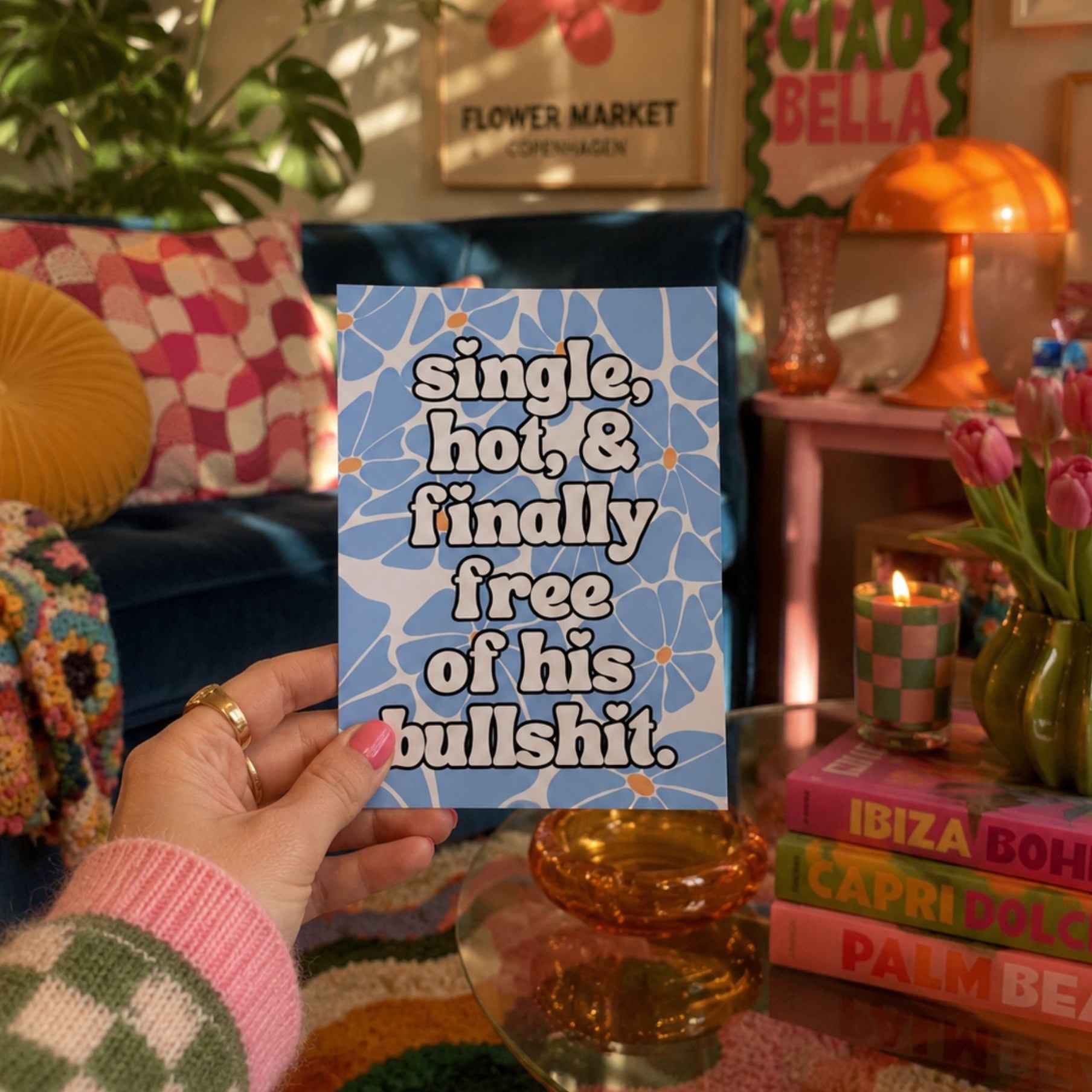A hand holds a blue card with a floral pattern and the text 'single, hoi, & finally free of his bullshit' in a cozy, retro-styled room with pink and orange decor.