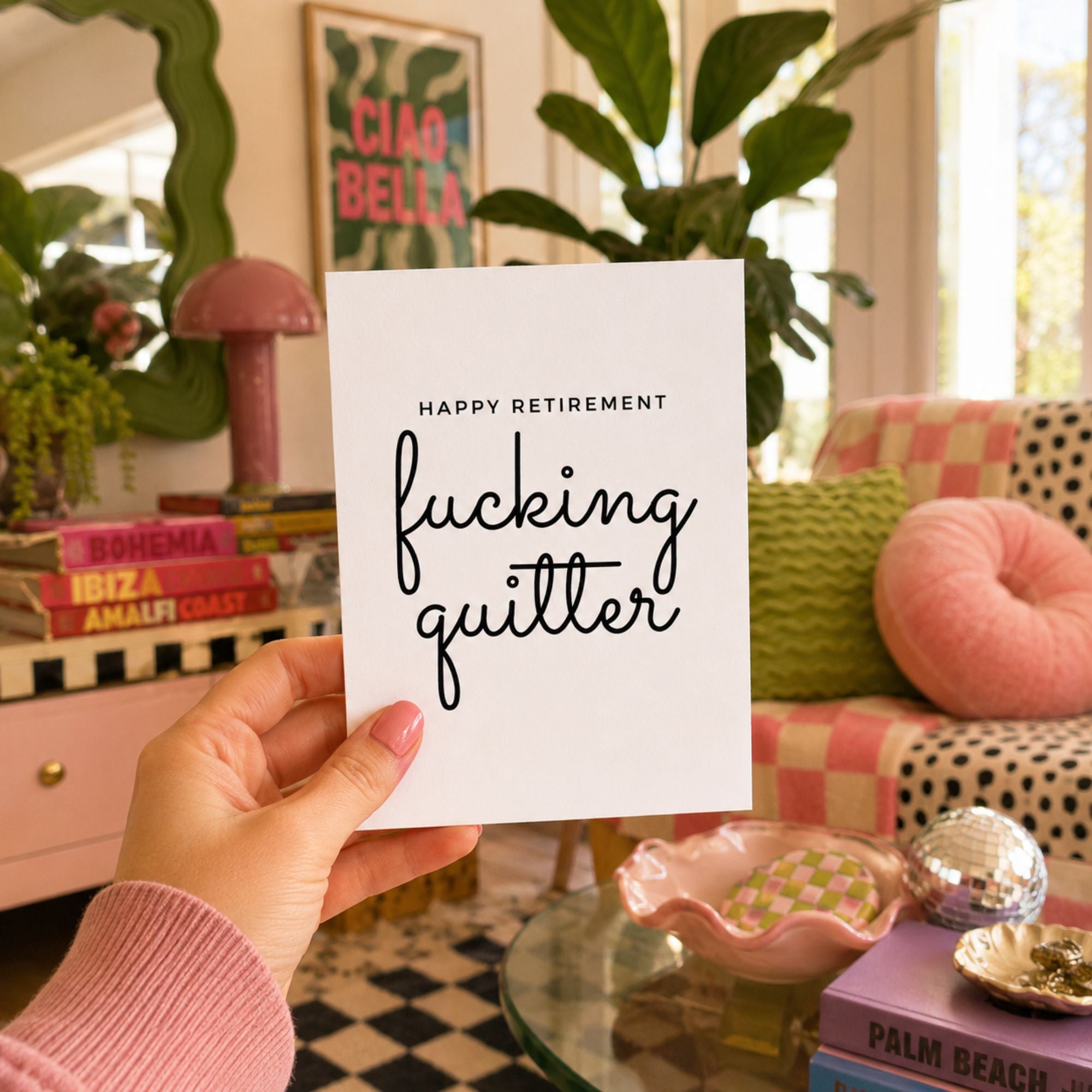 A hand holds a white greeting card that reads 'HAPPY RETIREMENT fucking quitter' in black text, set against a cozy, colorful living room background with a pink checkered sofa, green plants, and decorative items.