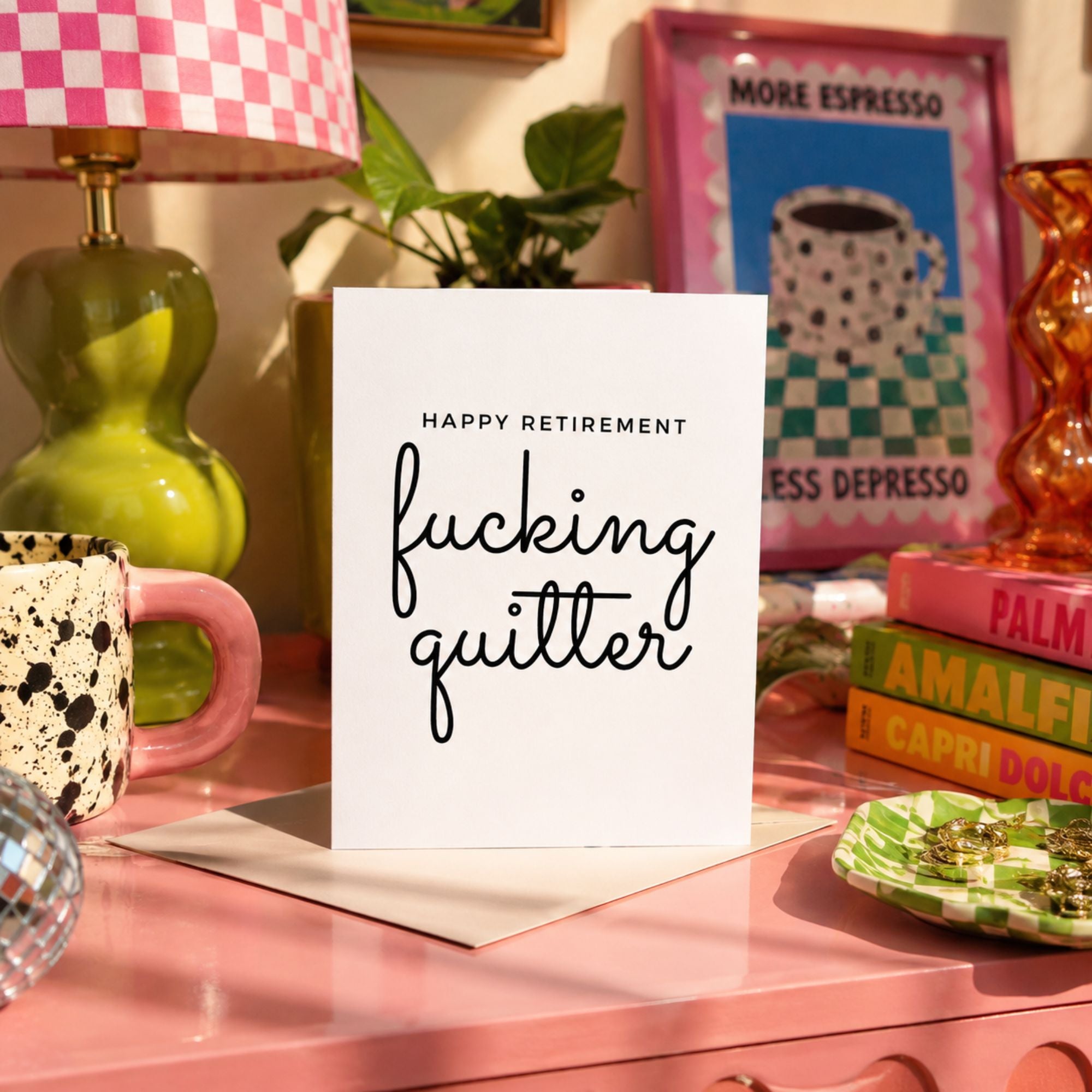 A white square greeting card with black text reading 'HAPPY RETIREMENT fucking quitter' stands on a pink tabletop in a styled room scene with a lamp, mug, books, and framed art in the background.