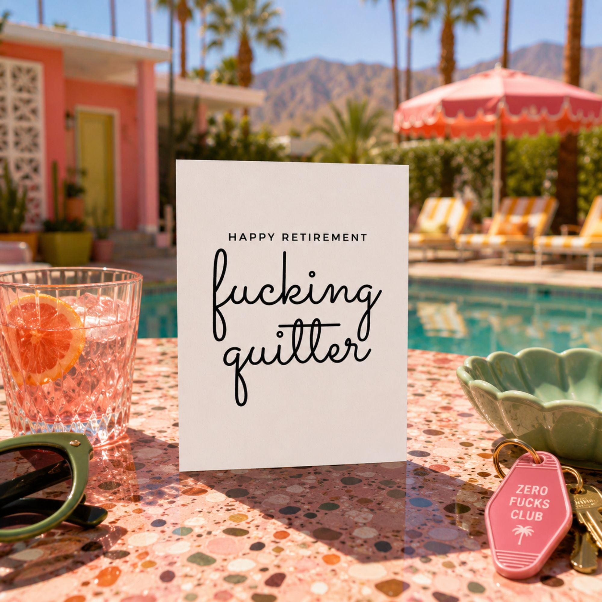 A white greeting card with black text reading 'HAPPY RETIREMENT' and 'fucking quitter' stands on a terrazzo poolside table. The scene includes a pink pool, lounge chairs, palm trees, sunglasses, a drink, and a pink keychain tag.