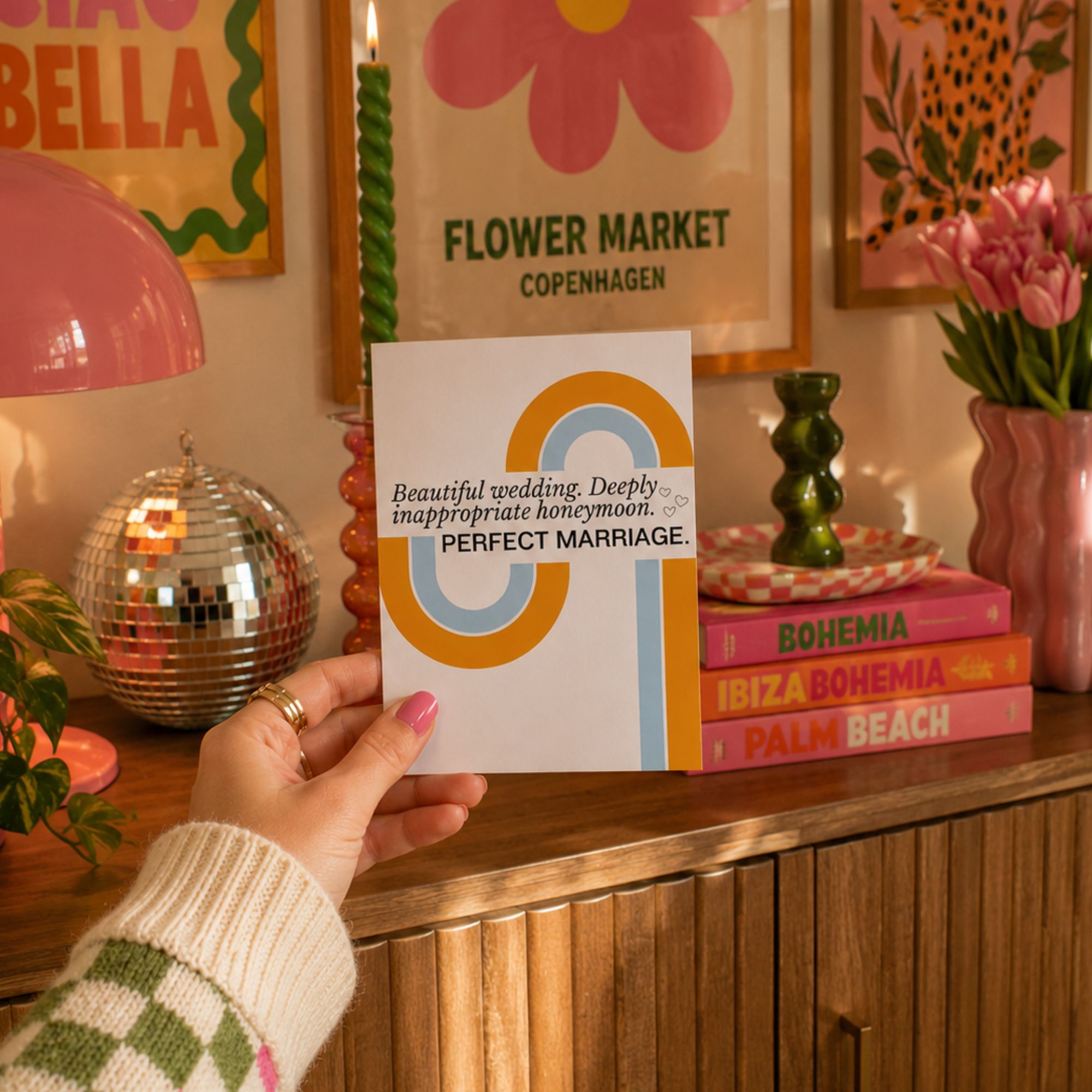 A hand holds a white wedding invitation card featuring a colorful abstract design and the text 'Beautiful wedding. Deeply inappropriate honeymoon.