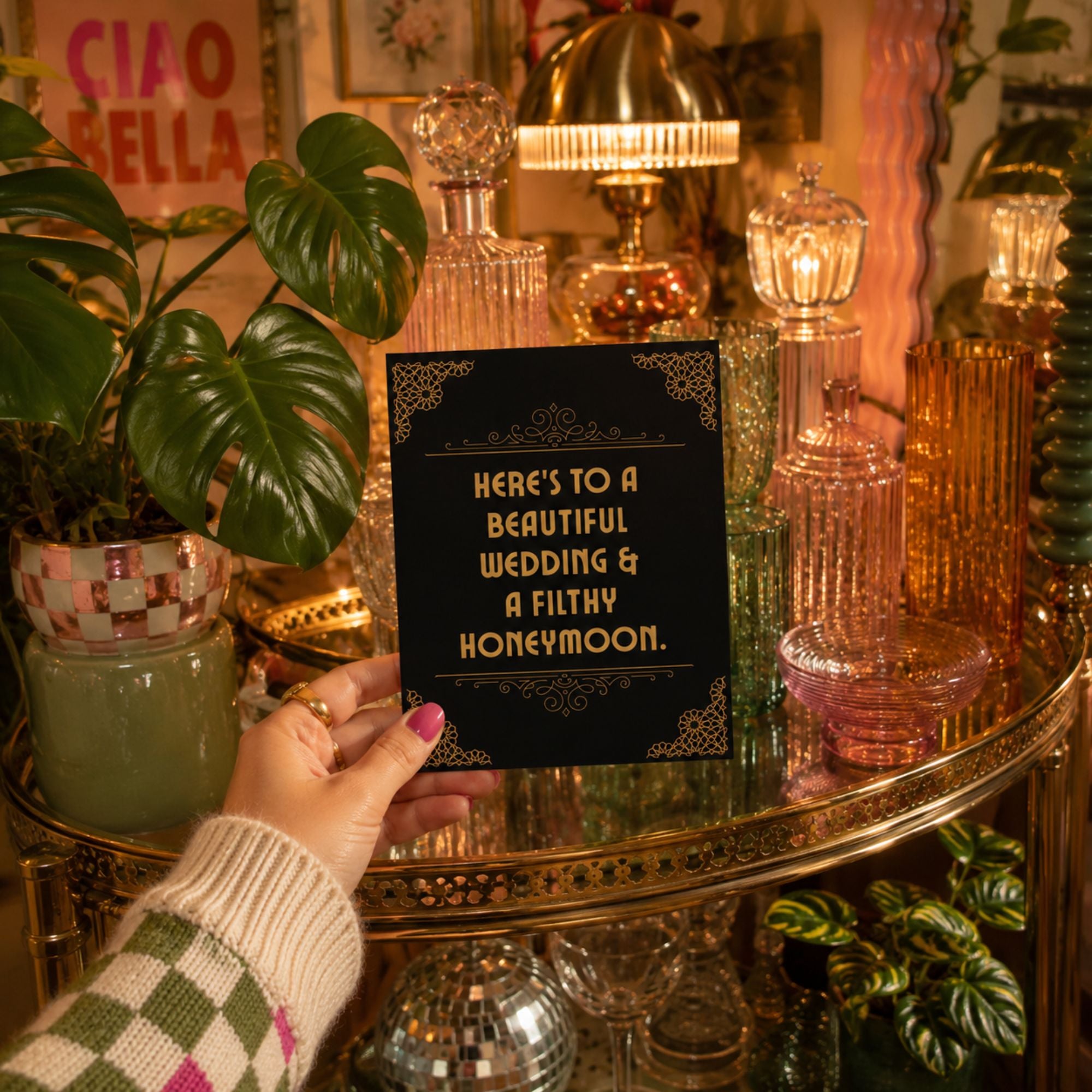 A hand in a green and pink knit sweater holds a black square sign with gold ornate borders and the text 'HERE'S TO A BEAUTIFUL WEDDING & A FILTHY HONEYMOON.' The sign is photographed on a reflective gold glass table surrounded by pink and amber glassware, plants, and warm lamps in a styled interior setting.