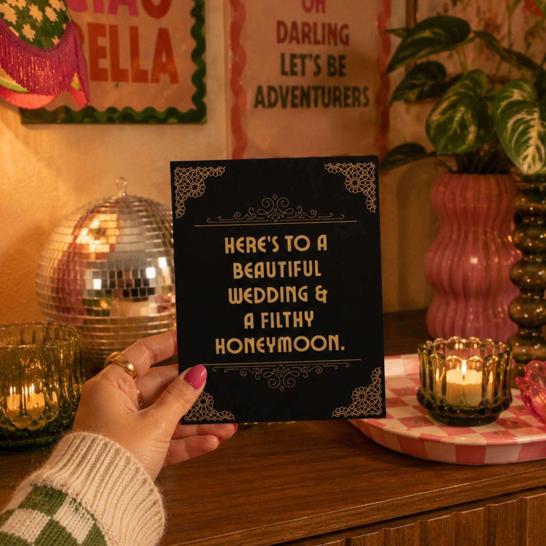 A hand holds a black wedding invitation card with gold ornate corner details and gold text reading 'HERE'S TO A BEAUTIFUL WEDDING & A FILTY HONEYMOON.' The card is shown in a warmly lit, retro-themed room setting with candles, a pink vase, and framed wall art in the background.