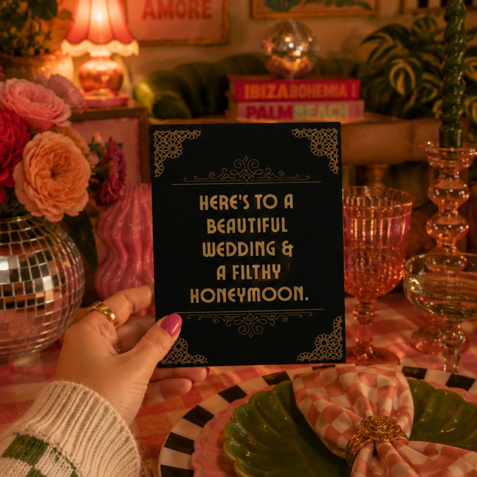 A hand holds a black wedding-themed card that reads 'HERE'S TO A BEAUTIFUL WEDDING & A FILTY HONEYMOON.' in gold lettering, set against a decorated table with pink checkered tablecloth, green plates, pink candles, and flowers.