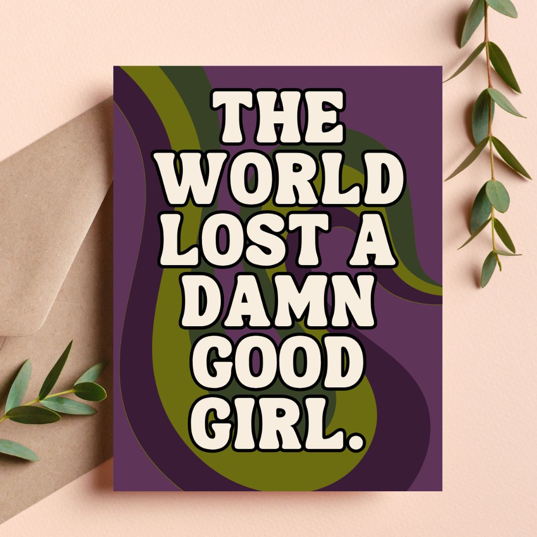 A greeting card with the text 'THE WORLD LOST A DAMN GOOD GIRL.' in bold white letters over a purple and olive green abstract background, styled on a beige surface with a kraft envelope and green leaves.