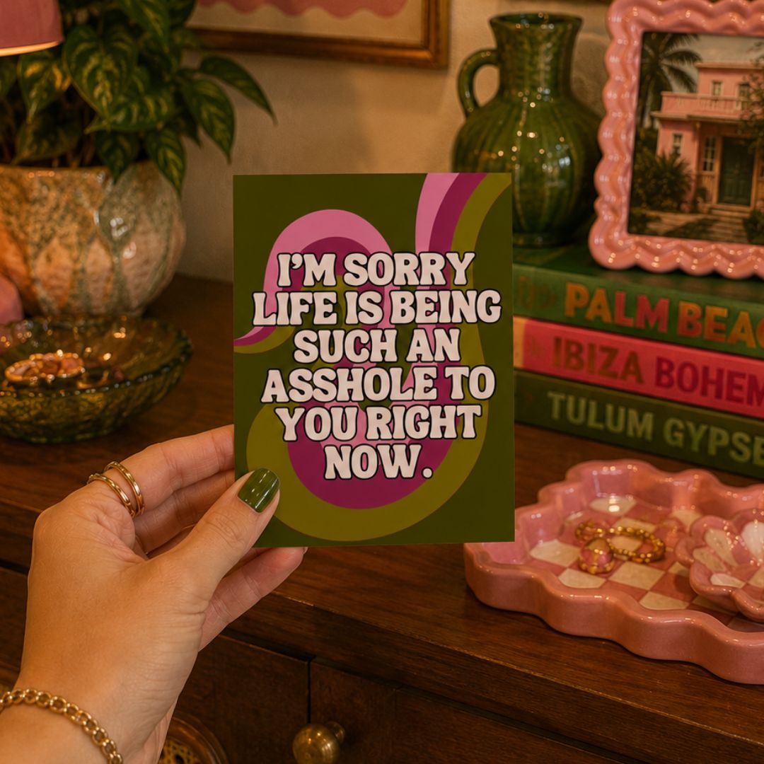 A hand holds a square printed card with a humorous quote that reads 'I'M SORRY LIFE IS BEING SUCH AN ASSHOLE TO YOU RIGHT NOW.' The card has a dark olive green background with a pink and purple abstract swoosh and white text.