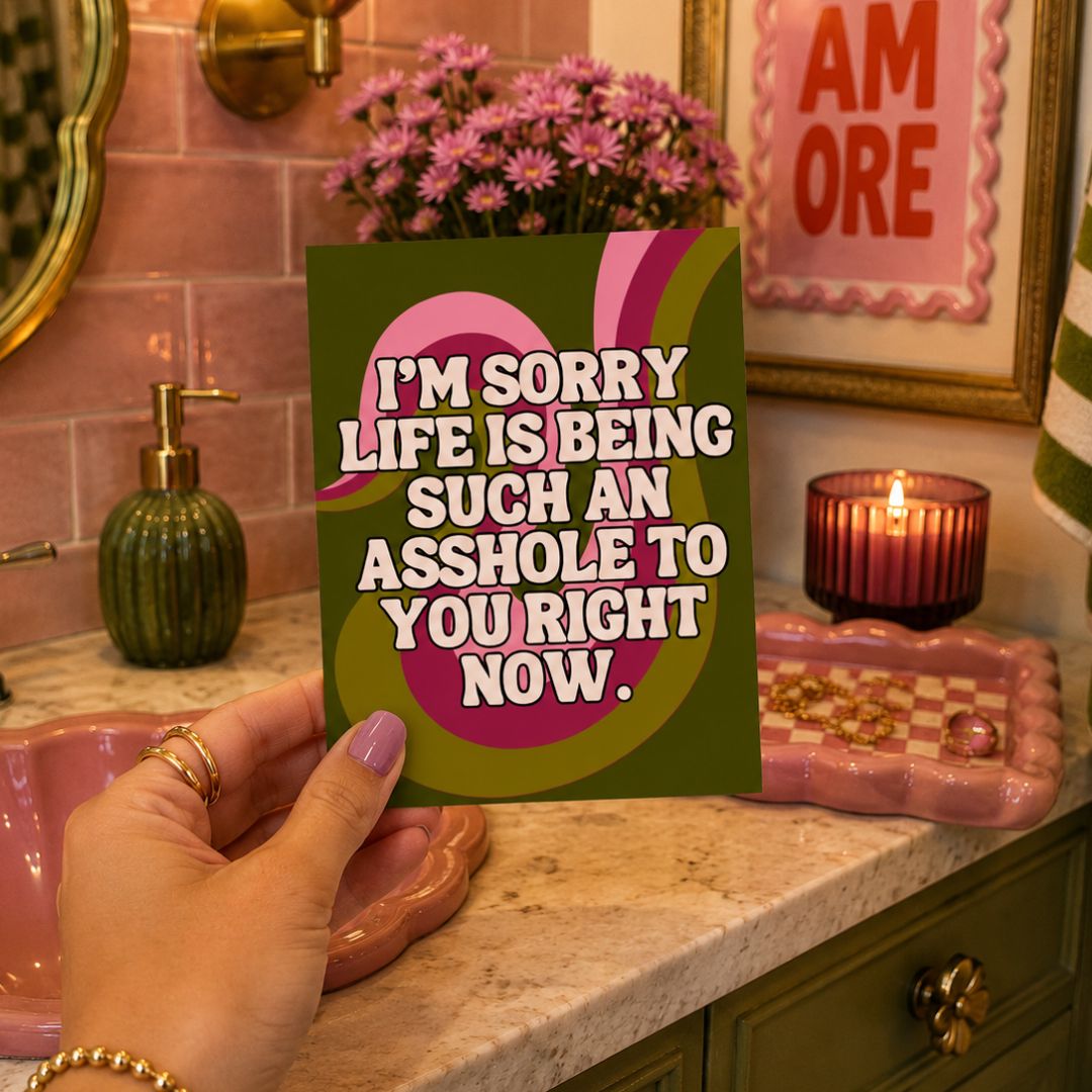 A hand holds a square greeting card with a humorous message, set against a cozy bathroom vanity background with pink tiles, flowers, and a lit candle.