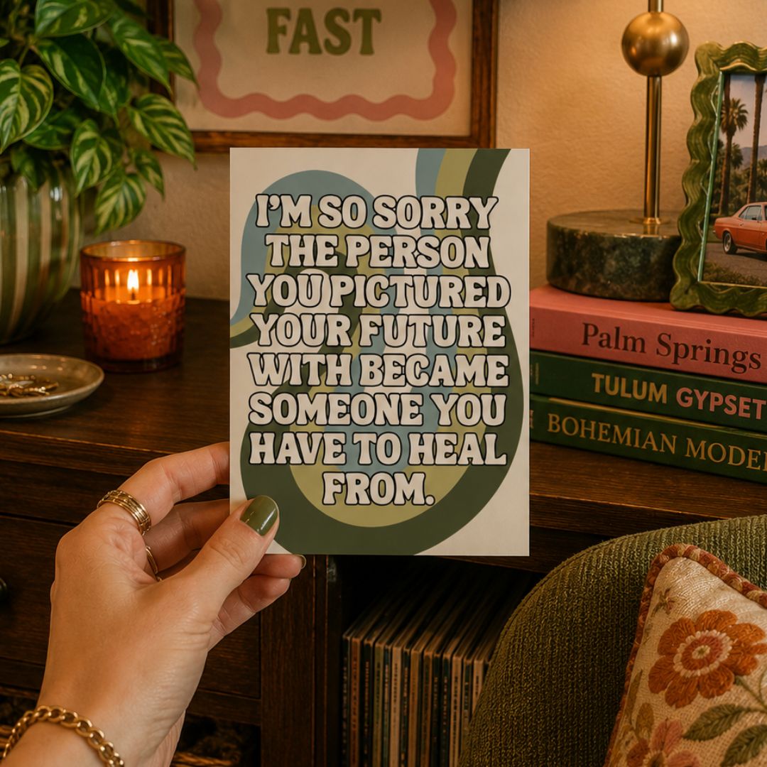 A hand holds a square art print with a humorous quote about healing. The print features a white background with a dark green and teal circular graphic and bold white text.