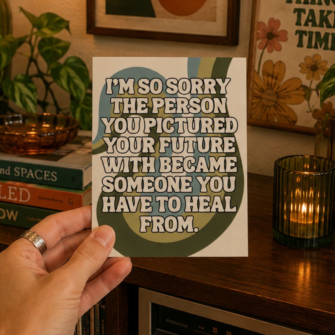 A hand holds a square card with a humorous quote in bold white text over a retro-style green, beige, and blue circular graphic. The background shows a cozy, warm-toned room with a wooden shelf, a lit candle, books, and framed art.