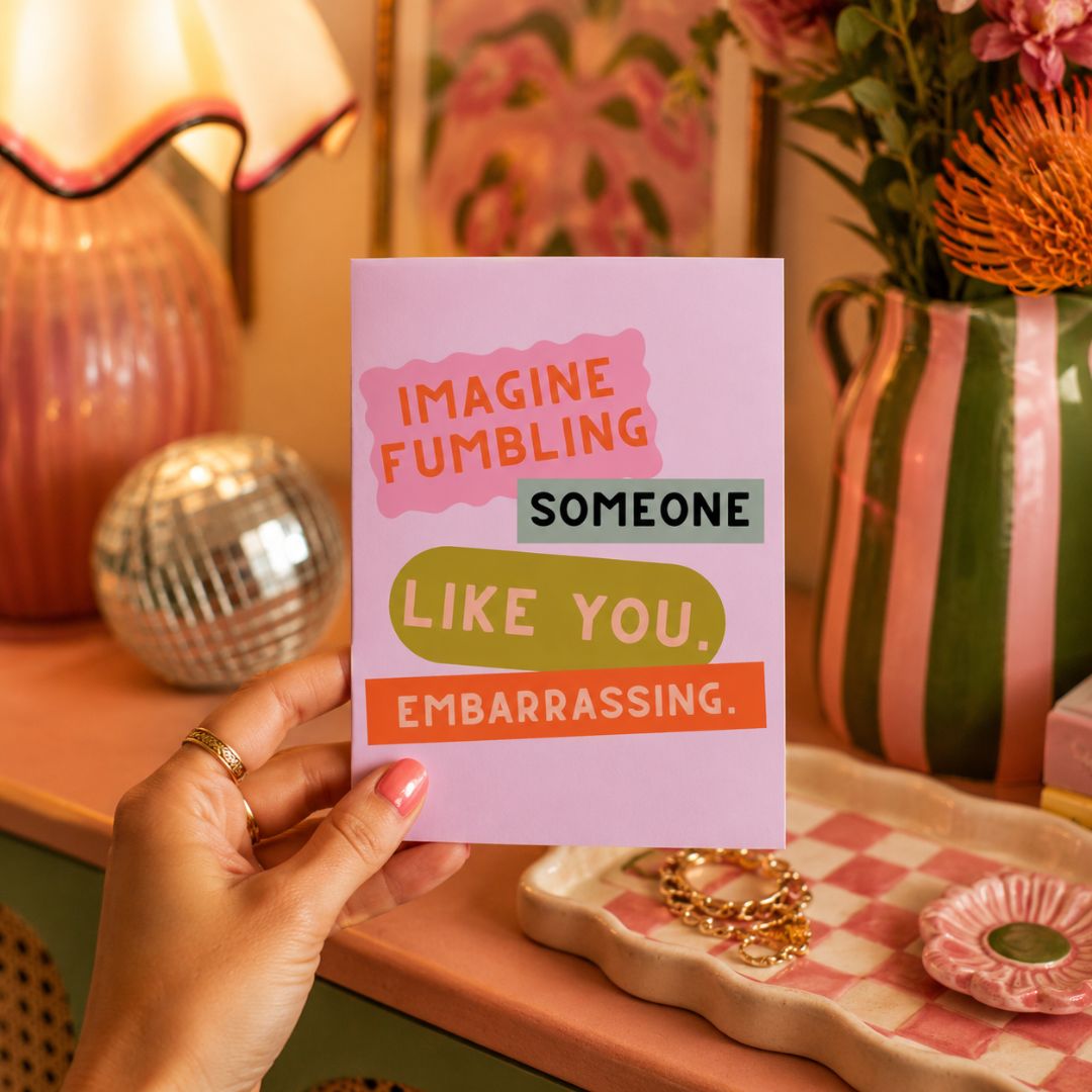 A hand holds a pink greeting card with the text 'IMAGINE FUMBING SOMEONE LIKE YOU, EMBARRASSING.' in colorful retro lettering.