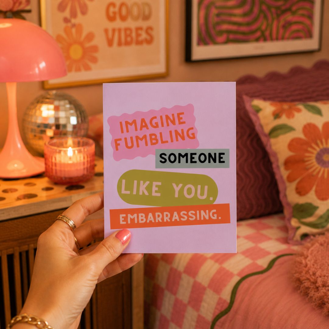A hand holds a pastel purple greeting card with colorful text that reads 'IMAGINE FUMBLING SOMEONE LIKE YOU. EMBARRASSING.