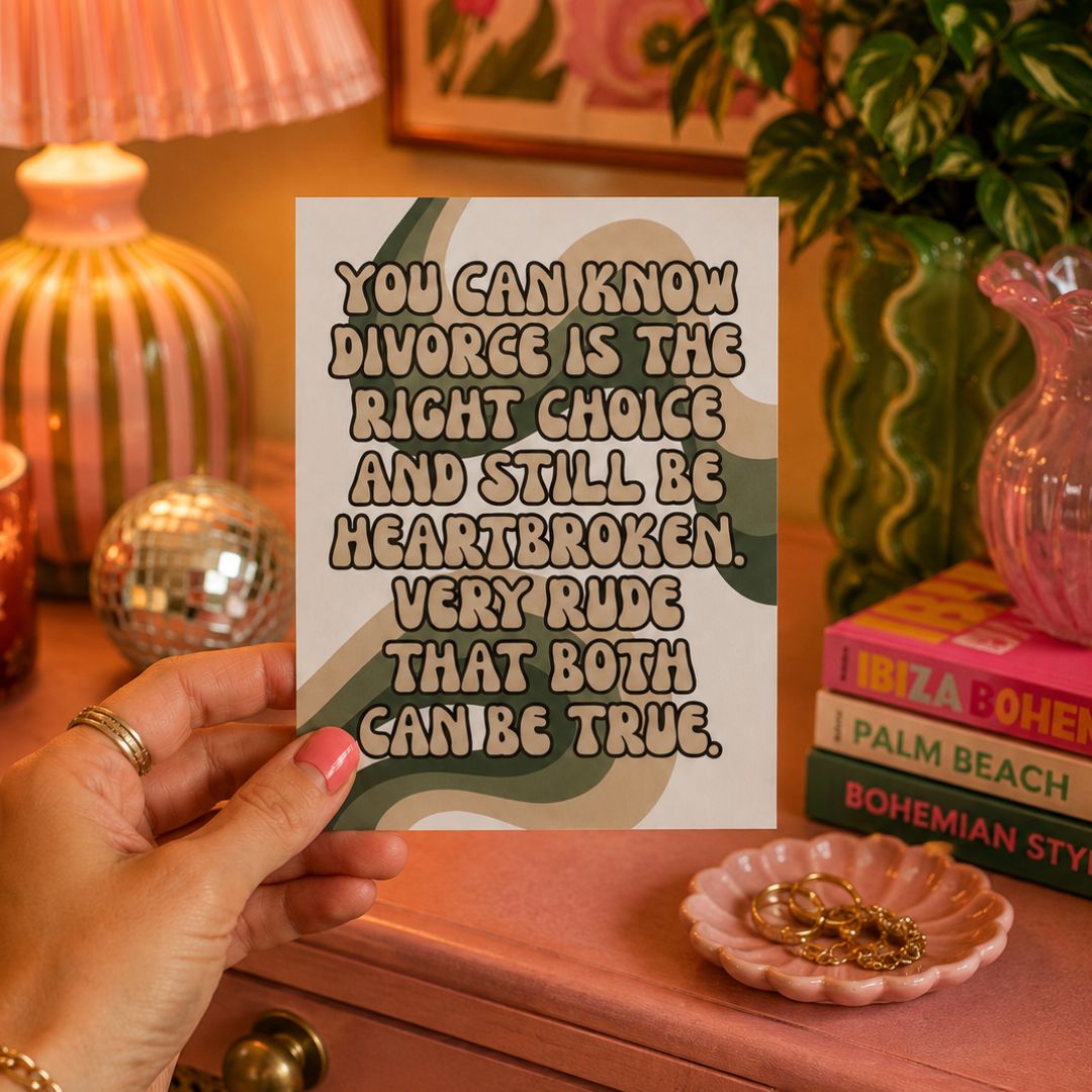 A hand holds a square art print featuring a humorous divorce quote in beige text over a green, beige, and gray retro wave pattern. The background is a cozy, warm-toned room with a pink dresser, a striped lamp, books, and jewelry.