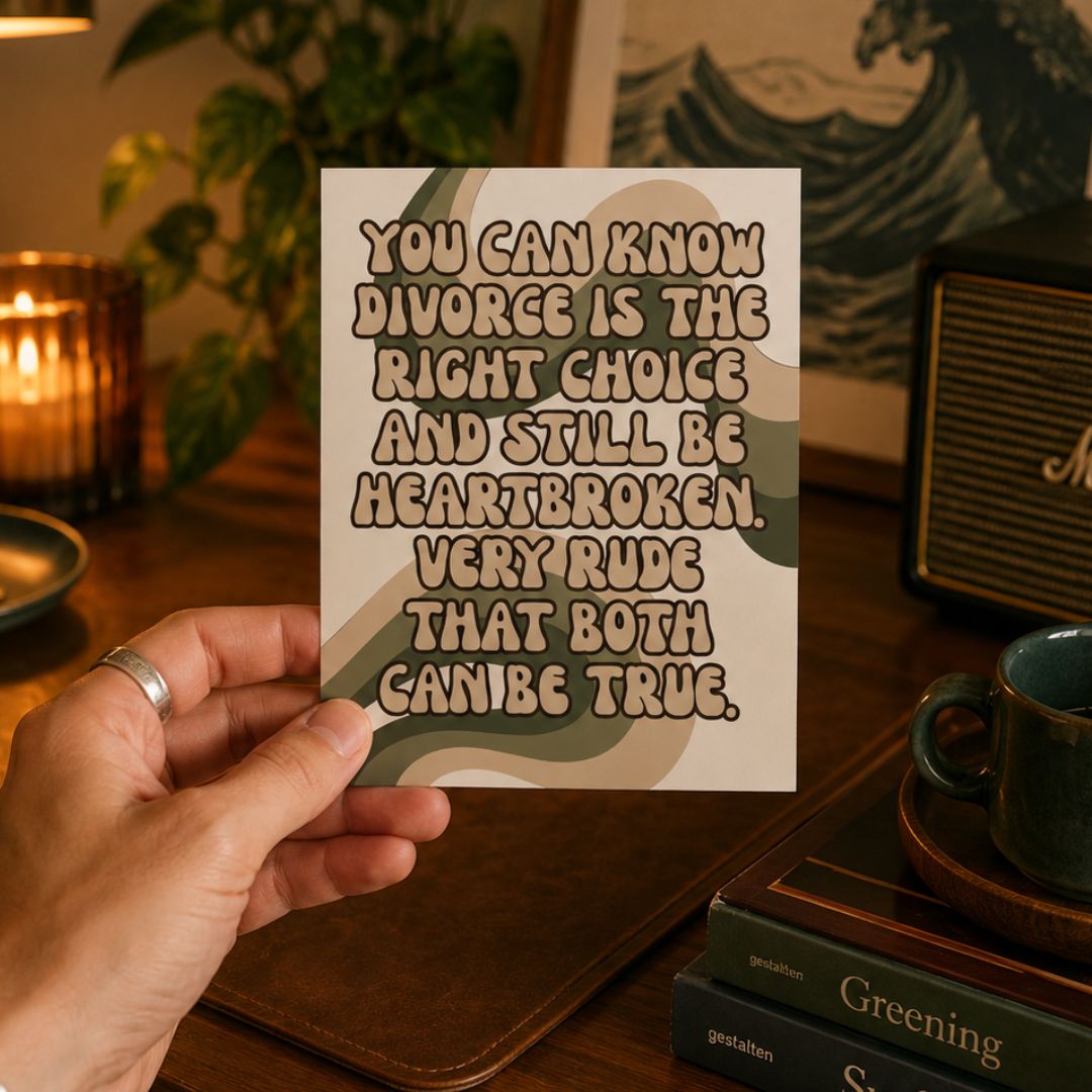 A hand holds a square art print with a humorous divorce quote in a warm, cozy room setting with a bookshelf, candle, and mug in the background.