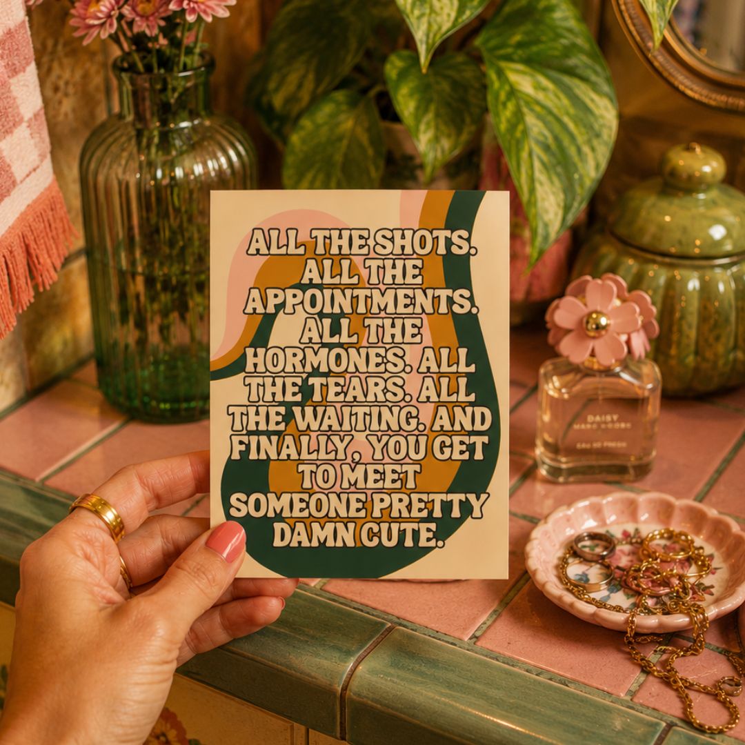 A hand holds a square card with a retro-style quote in cream, olive green, and mustard yellow. The card is set against a styled tabletop scene with a green glass vase, pink flowers, a perfume bottle, and a pink jewelry dish.