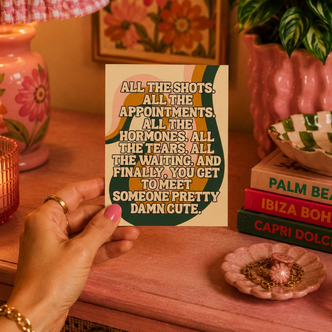 A hand holds a retro-style humorous card with a quote, set against a warm, bohemian-themed background with pink ceramic vases, books, and a pink table.