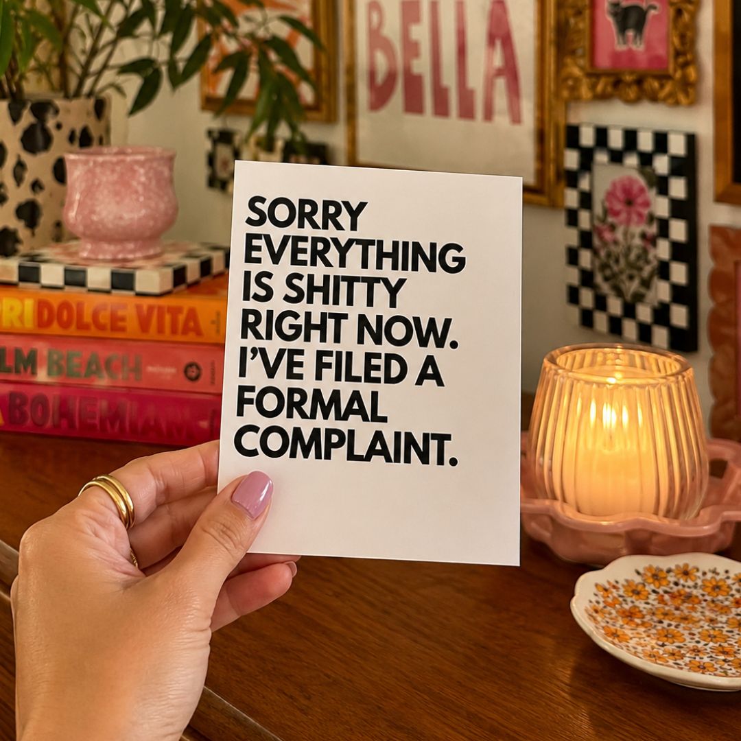 A hand holds a white rectangular card with bold black text reading, 'SORRY EVERYTHING IS SHITTY RIGHT NOW. I'VE FILED A FORMAL COMPLAINT.