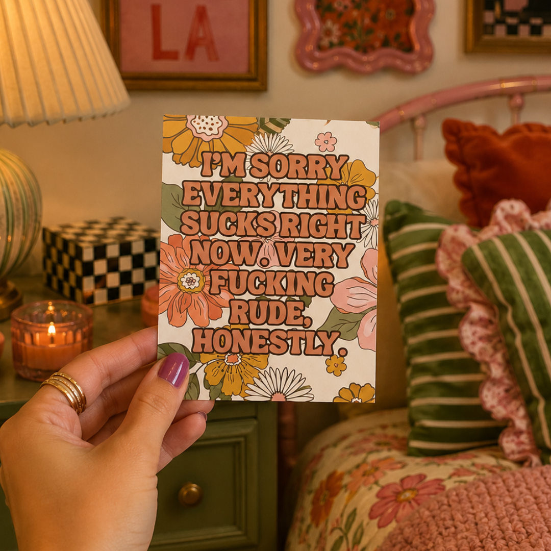 A hand holds a square floral card with bold retro text reading, 'I'M SORRY EVERYTHING SUCKS RIGHT NOW. VERY FUCKING RUDE, HONESTLY.
