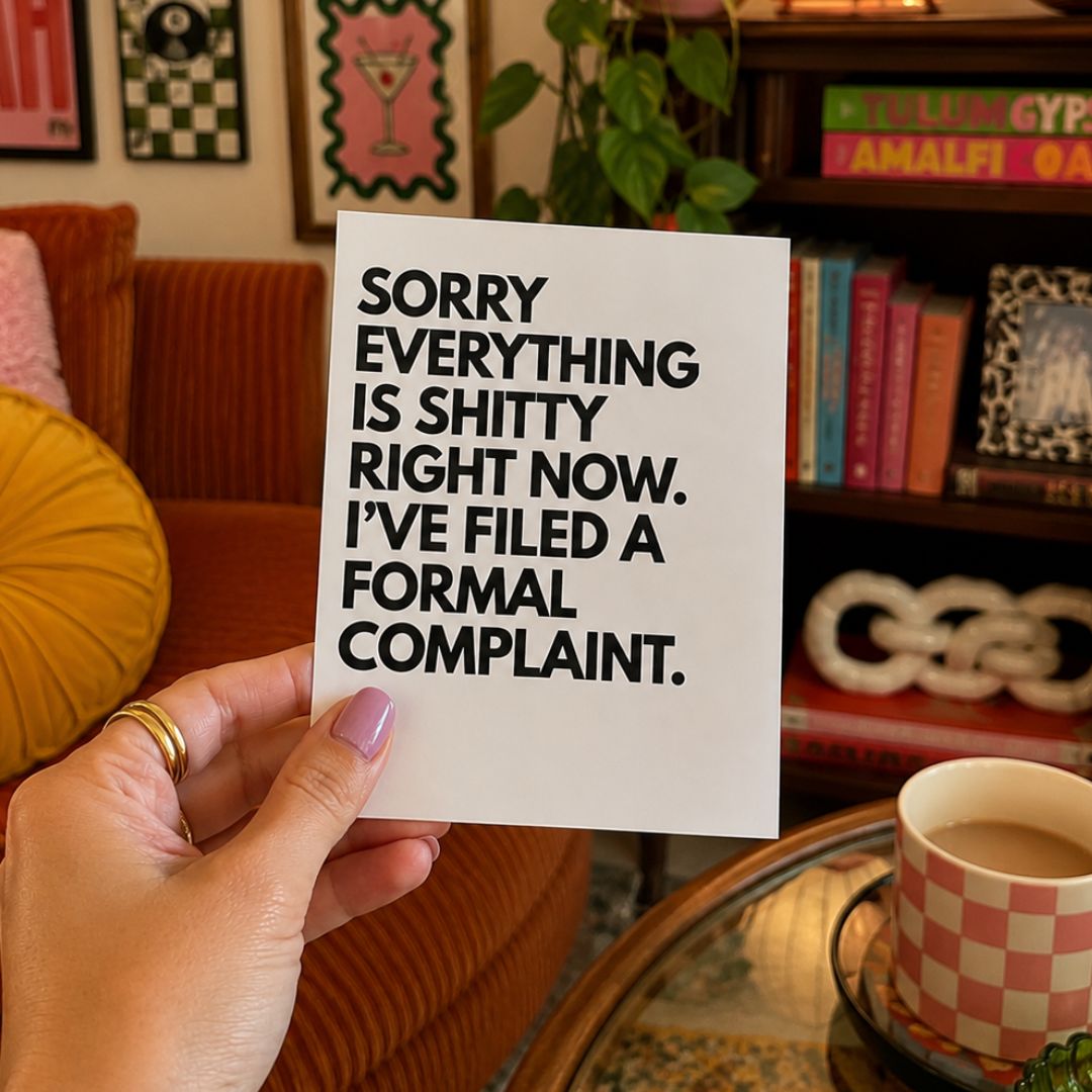 A hand holds a white square card with bold black text reading, 'SORRY EVERYTHING IS SHITTY RIGHT NOW. I'VE FILED A FORMAL COMPLAINT.
