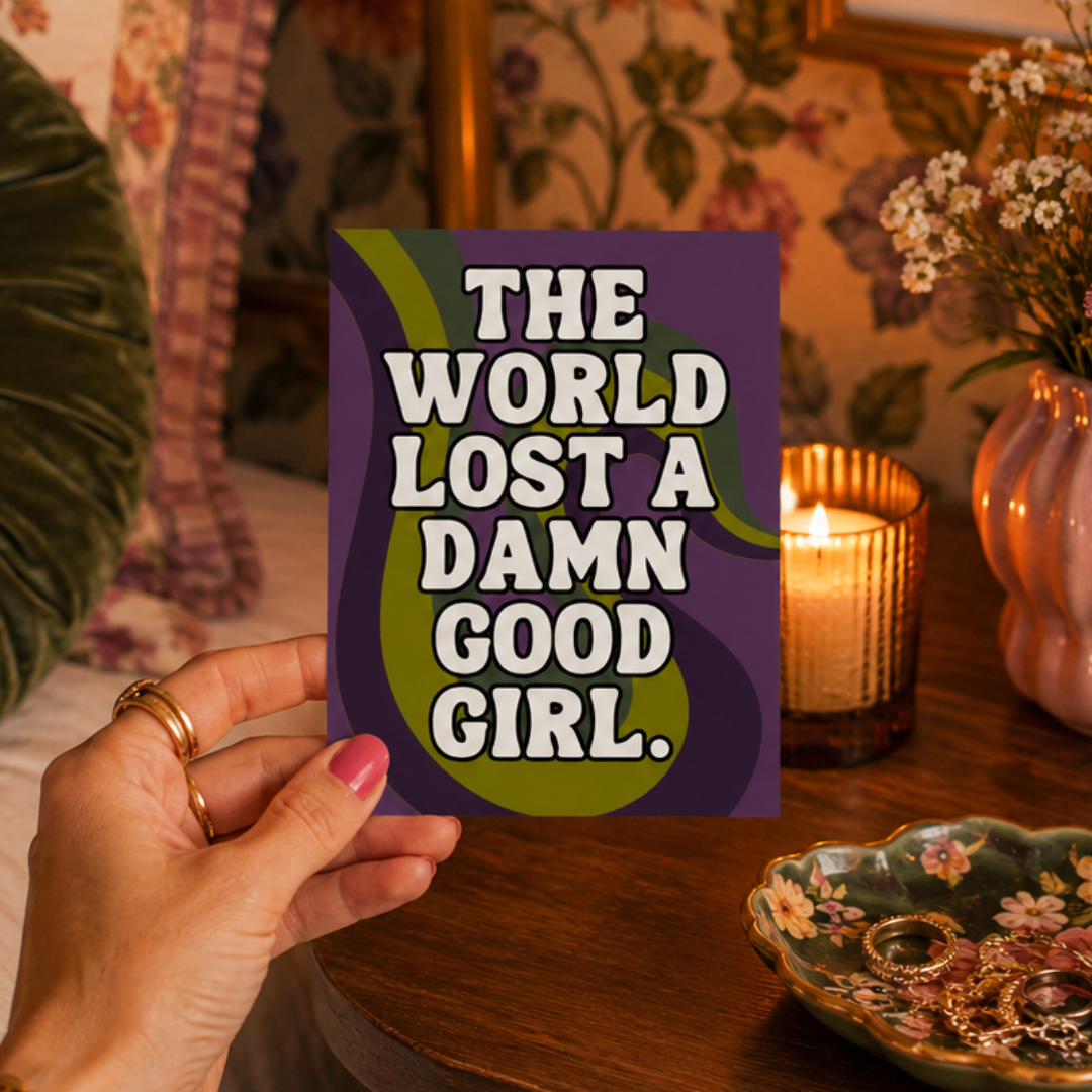 A hand holds a square art print with the text 'THE WORLD LOST A DAMN GOOD GIRL.' in white letters over a purple and olive green background.