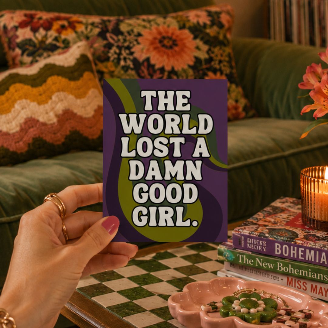 A hand holds a square art print with the text 'THE WORLD LOST A DAMN GOOD GIRL.' in white letters over a purple and olive green background.