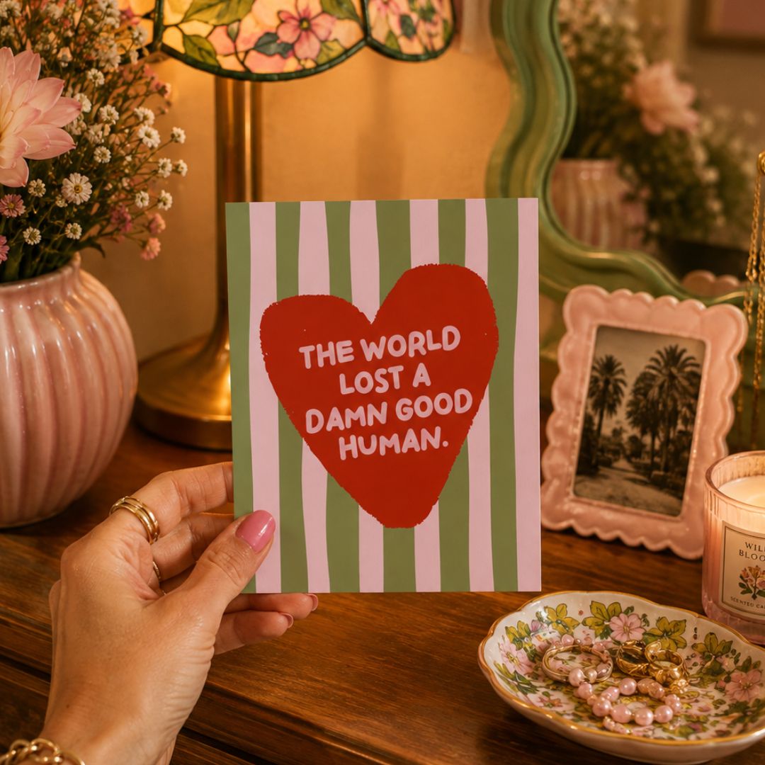 A hand holds a square greeting card with green and white vertical stripes and a large red heart in the center containing the text 'THE WORLD LOST A DAMN GOOD HUMAN.' The card is photographed in a cozy, styled room setting with a pink ribbed vase of flowers, a lamp, a framed palm tree photo, and a floral dish on a wooden table.