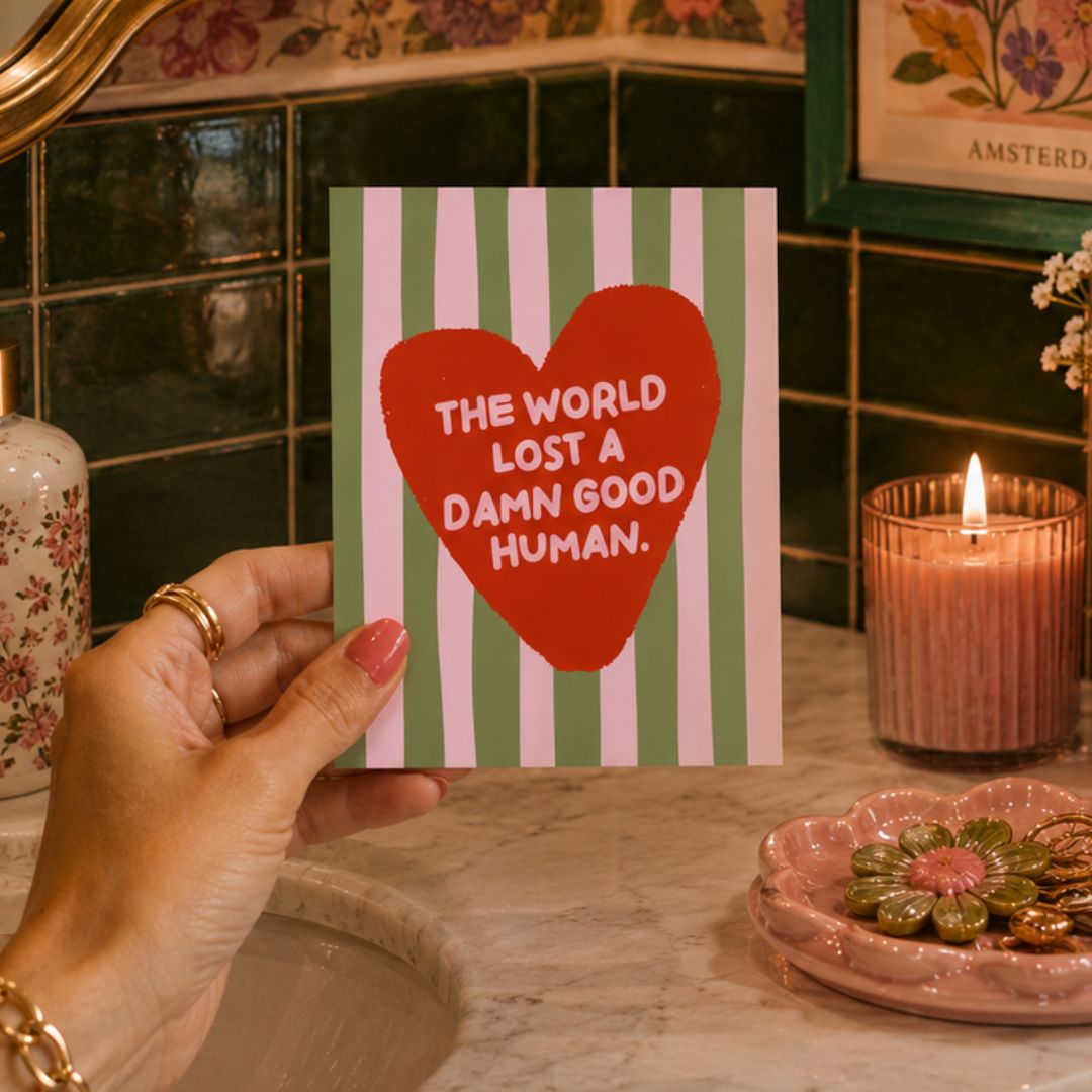 A hand holds a greeting card with a red heart and the text 'THE WORLD LOST A DAMN GOOD HUMAN.' against a background of a bathroom sink, green tiles, a lit candle, and a pink flower-shaped dish.