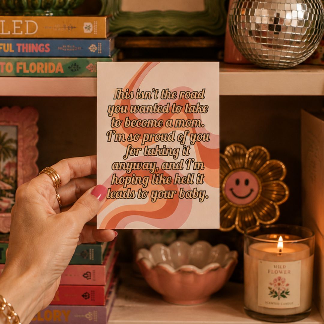 A hand holds a square greeting card with a humorous quote for a new mom. The card features a white background with retro wavy stripes in orange and pink, and the text is written in a brown cursive font.