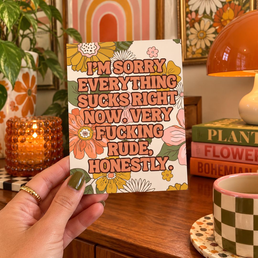 A hand holds a square card with a floral border and bold text that reads, 'I'M SORRY EVERYTHING SUCKS RIGHT NOW. VERY FUCKING RUDE, HONESTLY.
