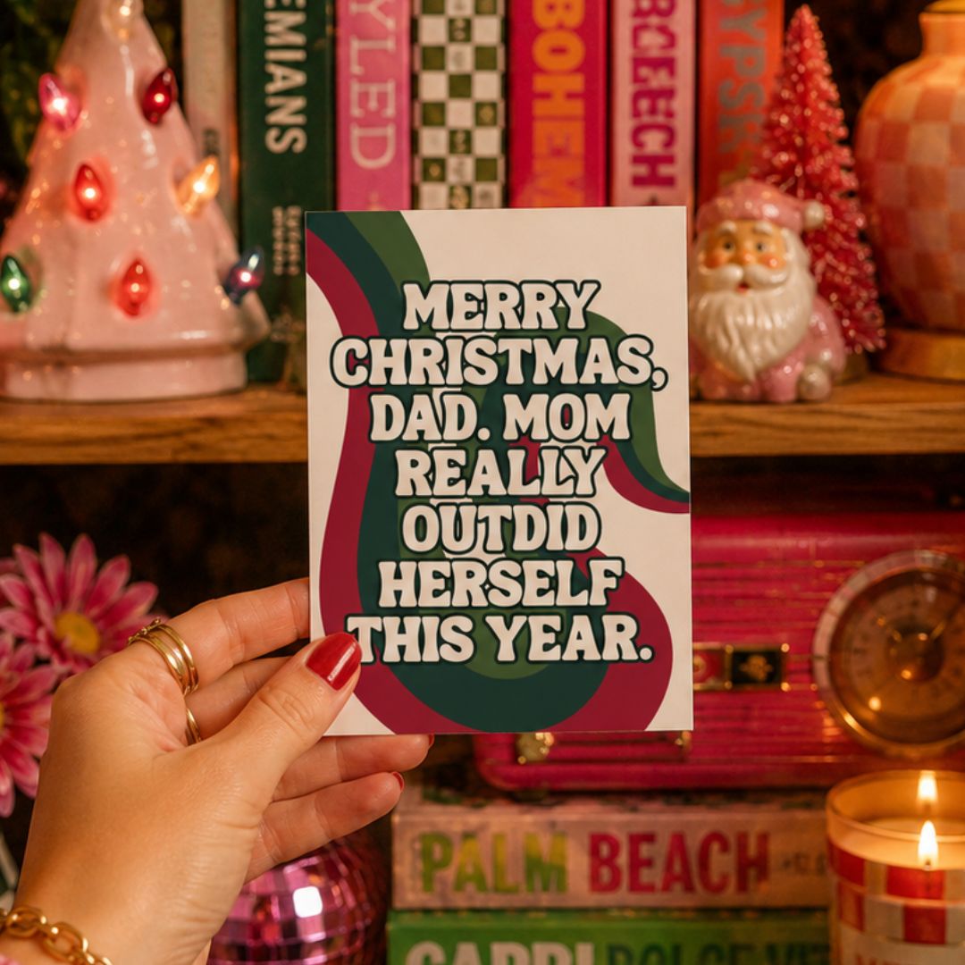 A hand holds a white Christmas greeting card with a humorous quote, set against a cozy, festive background of bookshelves and holiday decorations.