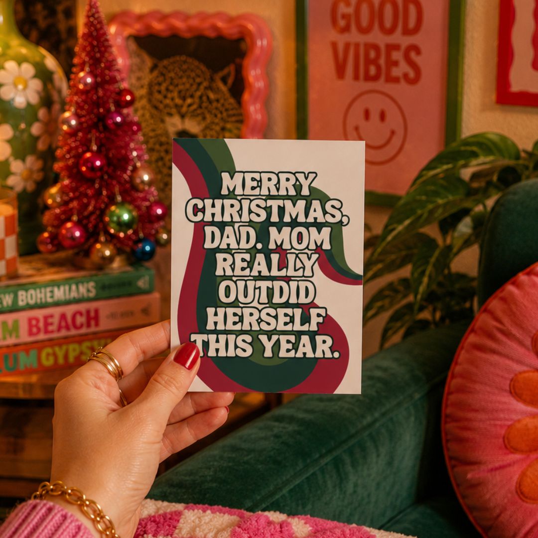 A hand with red nail polish holds a white Christmas greeting card with a humorous quote for Dad. The card features a dark green and burgundy abstract design.